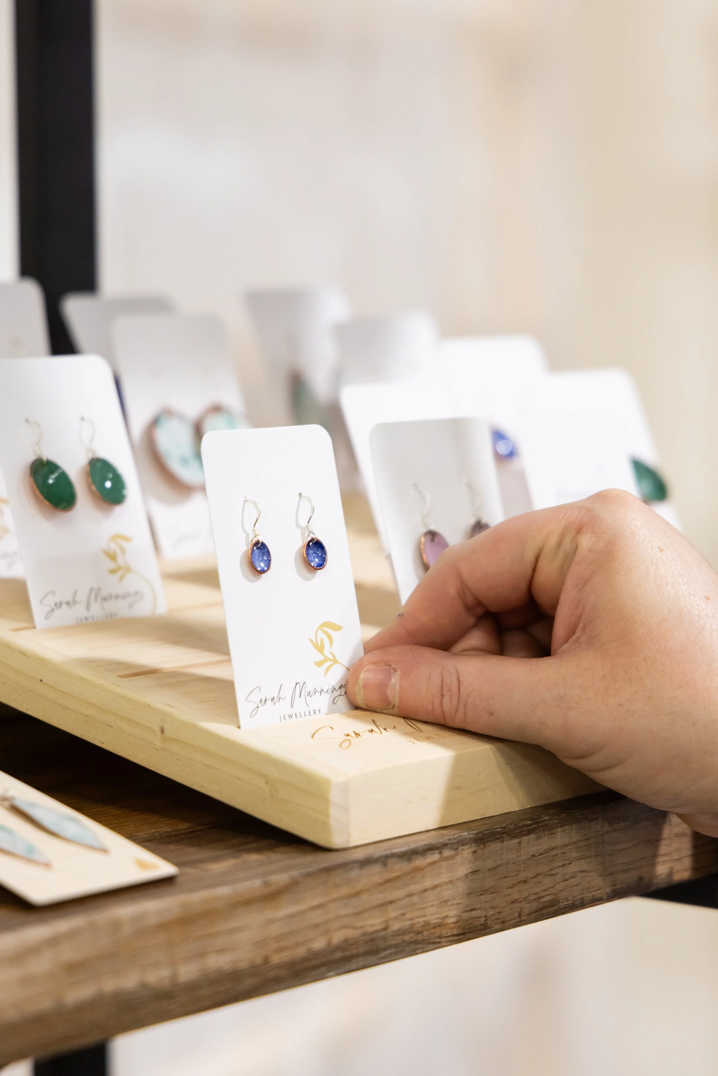 Person holding a blue teardrop-shaped earring in front of a display of various colored earrings on a wooden table.