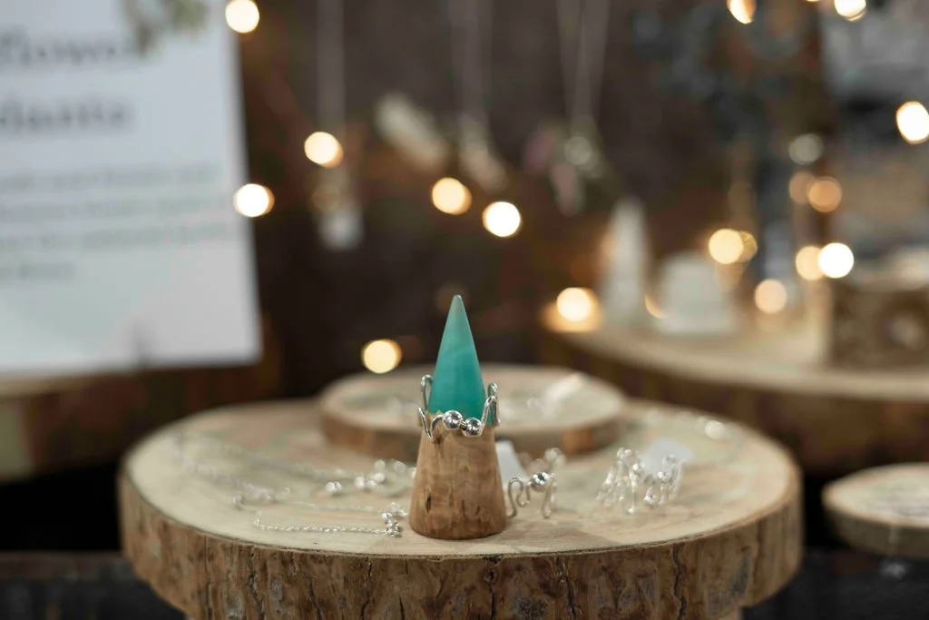 Decorative jewelry display on a wooden slab with a glass cone as a ring holder, necklaces, and earrings, with blurred string lights in the background.
