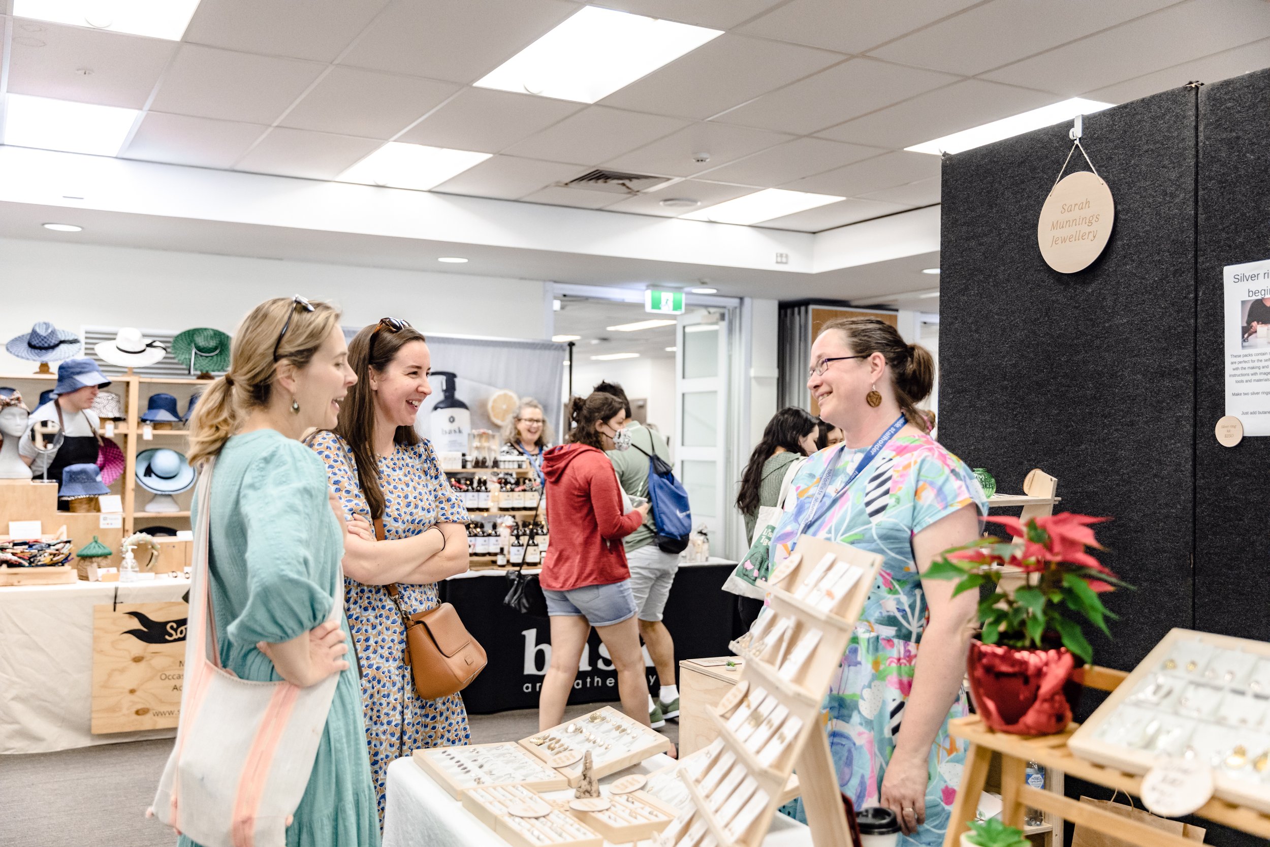 Three women smiling and talking at a craft fair, with jewelry and accessories on display behind them.