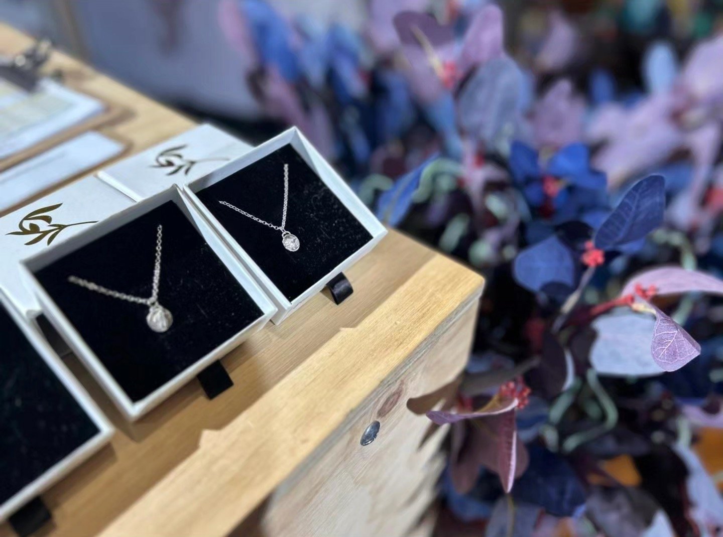Two boxed necklaces displayed on a wooden surface, with a background of purple and green foliage plants.