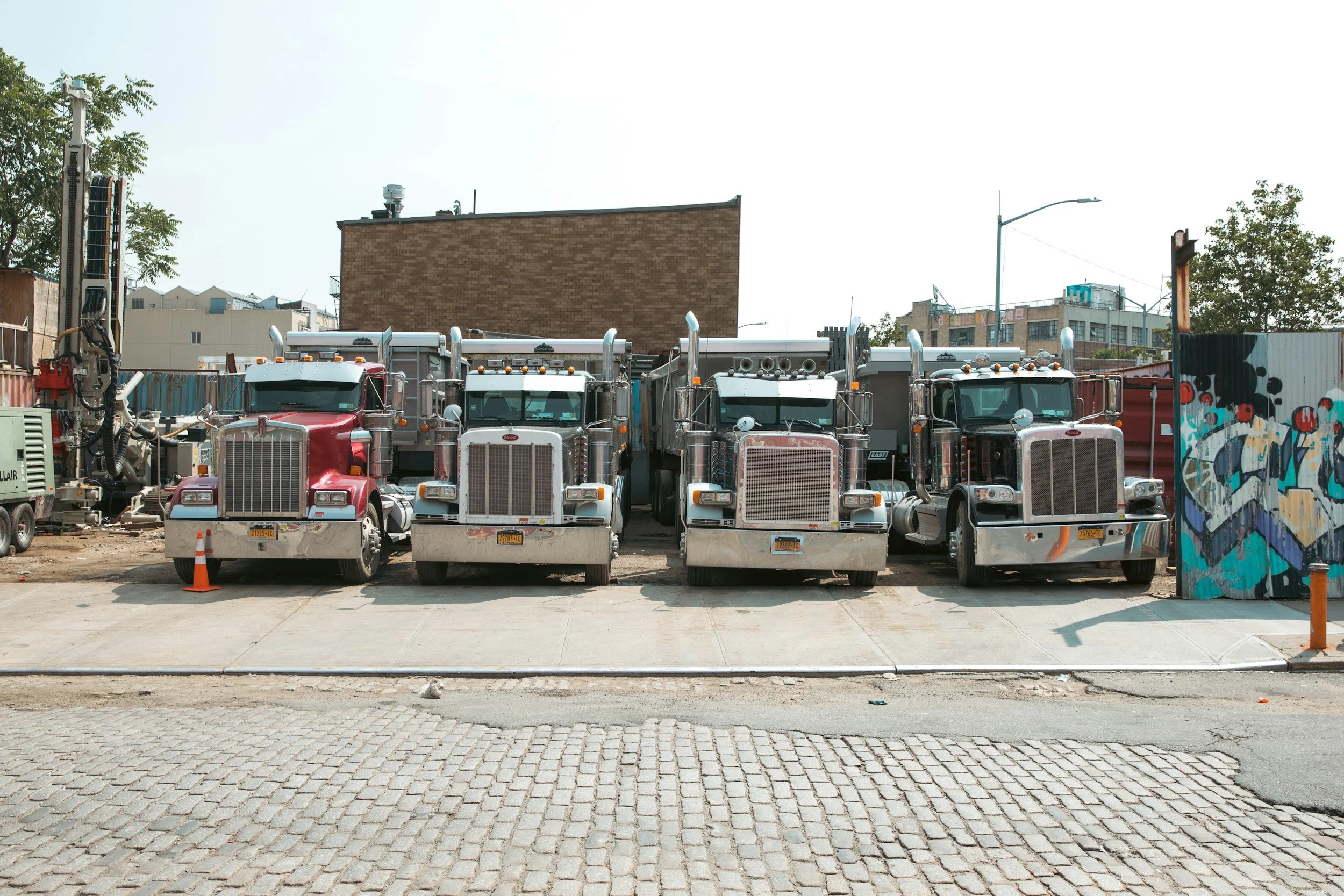 Four semi-trucks parked side by side on a sidewalk in an urban area, with a colorful graffiti wall on the right and multi-story buildings in the background.