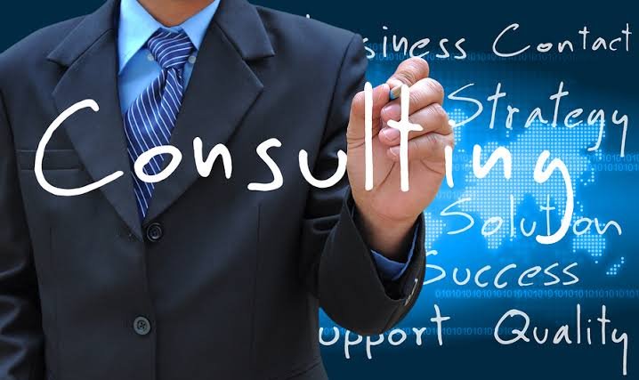 Business person in a suit writing the word 'Consulting' on a transparent screen with business-related terms in the background.