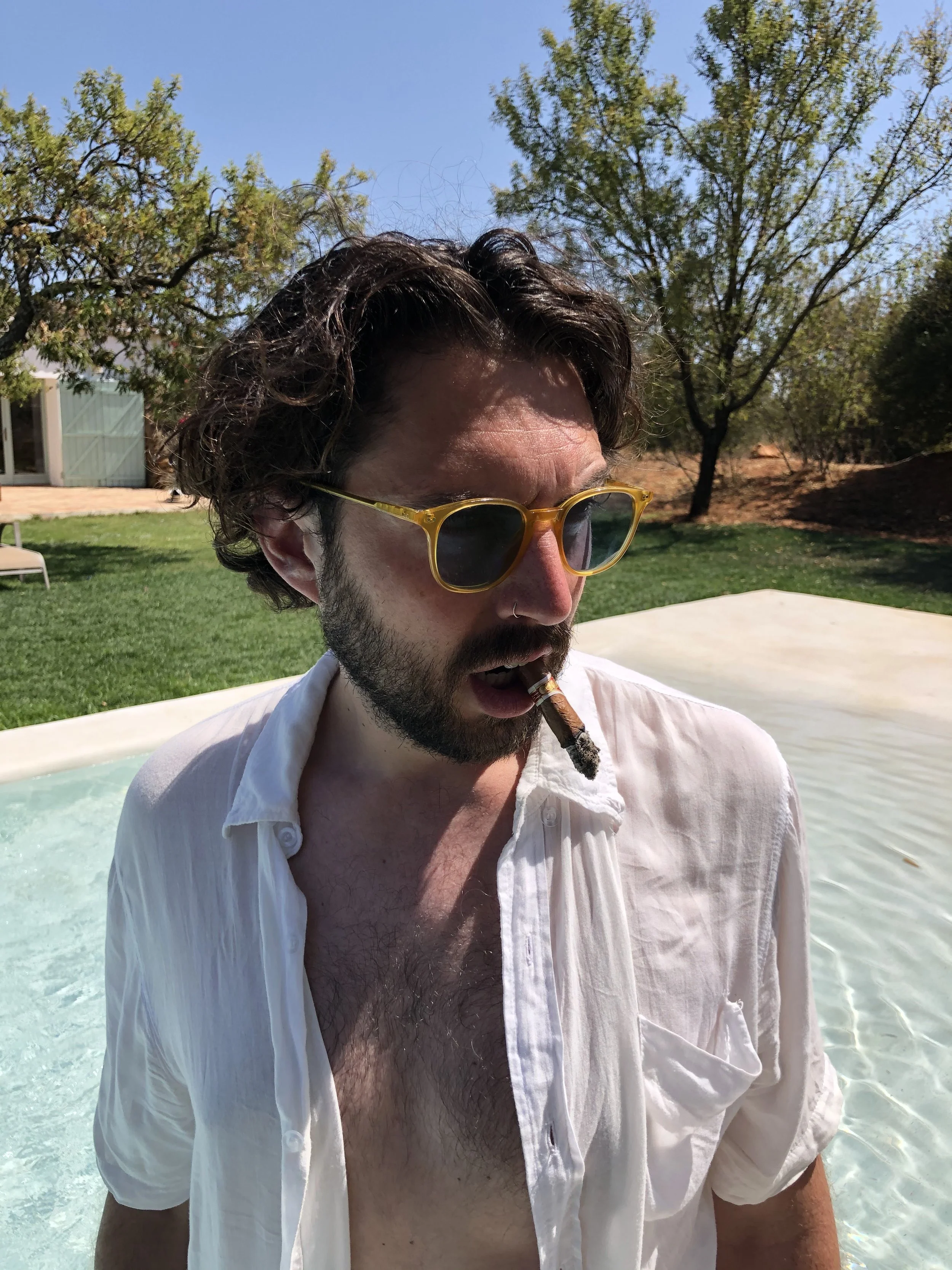 Man wearing sunglasses and a white shirt standing in a pool somewhere in Portugal with a cigar in his mouth, trees and grass in the background.