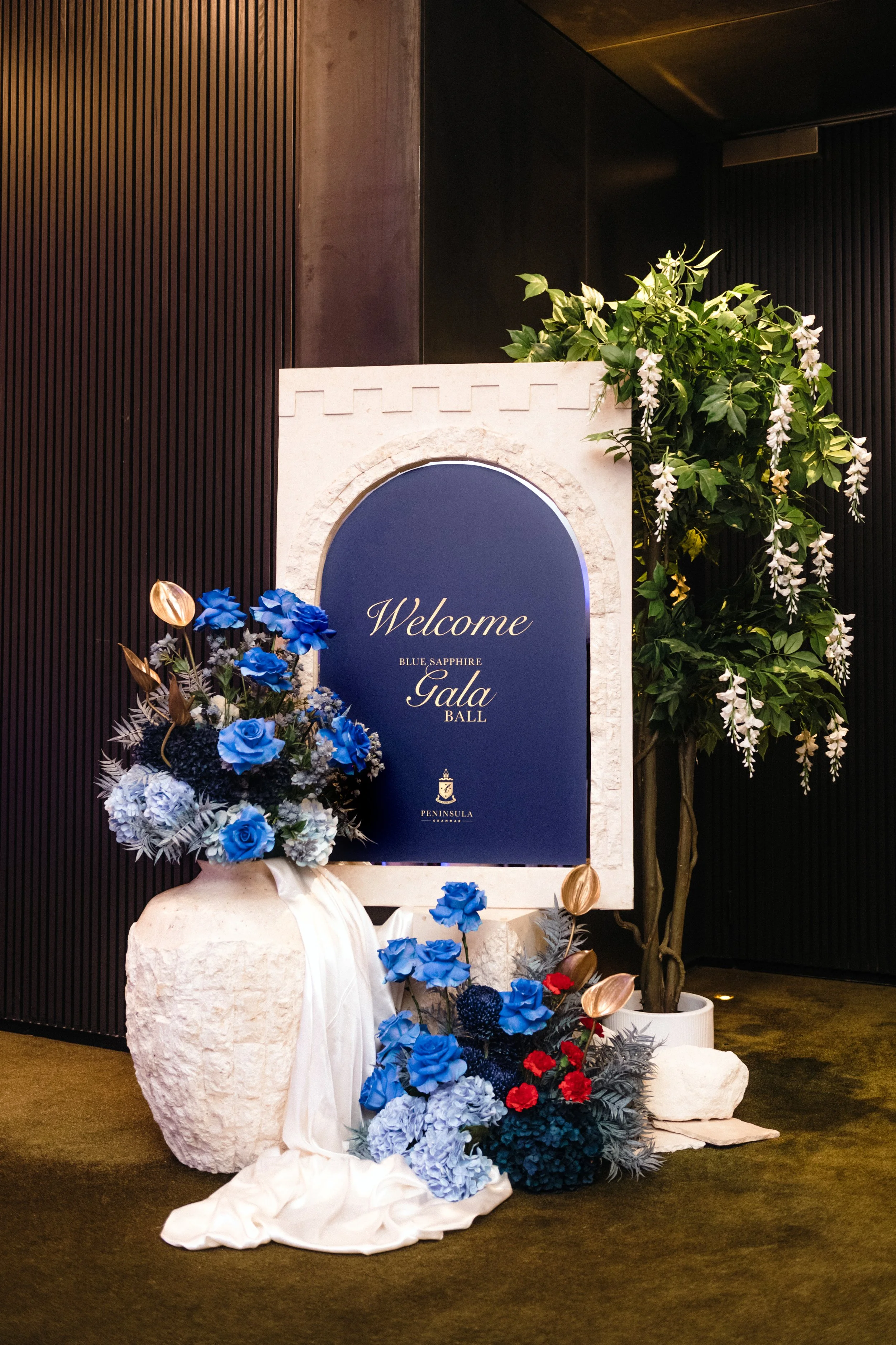 Modern‑lux corporate event styling by Poppy Culture at RACV Cape Schanck Resort, featuring a deep‑blue “Welcome Blue Sapphire Gala Ball” sign framed in a white architectural arch. Lush florals in blue roses, pale hydrangeas, red accents, gold foliage