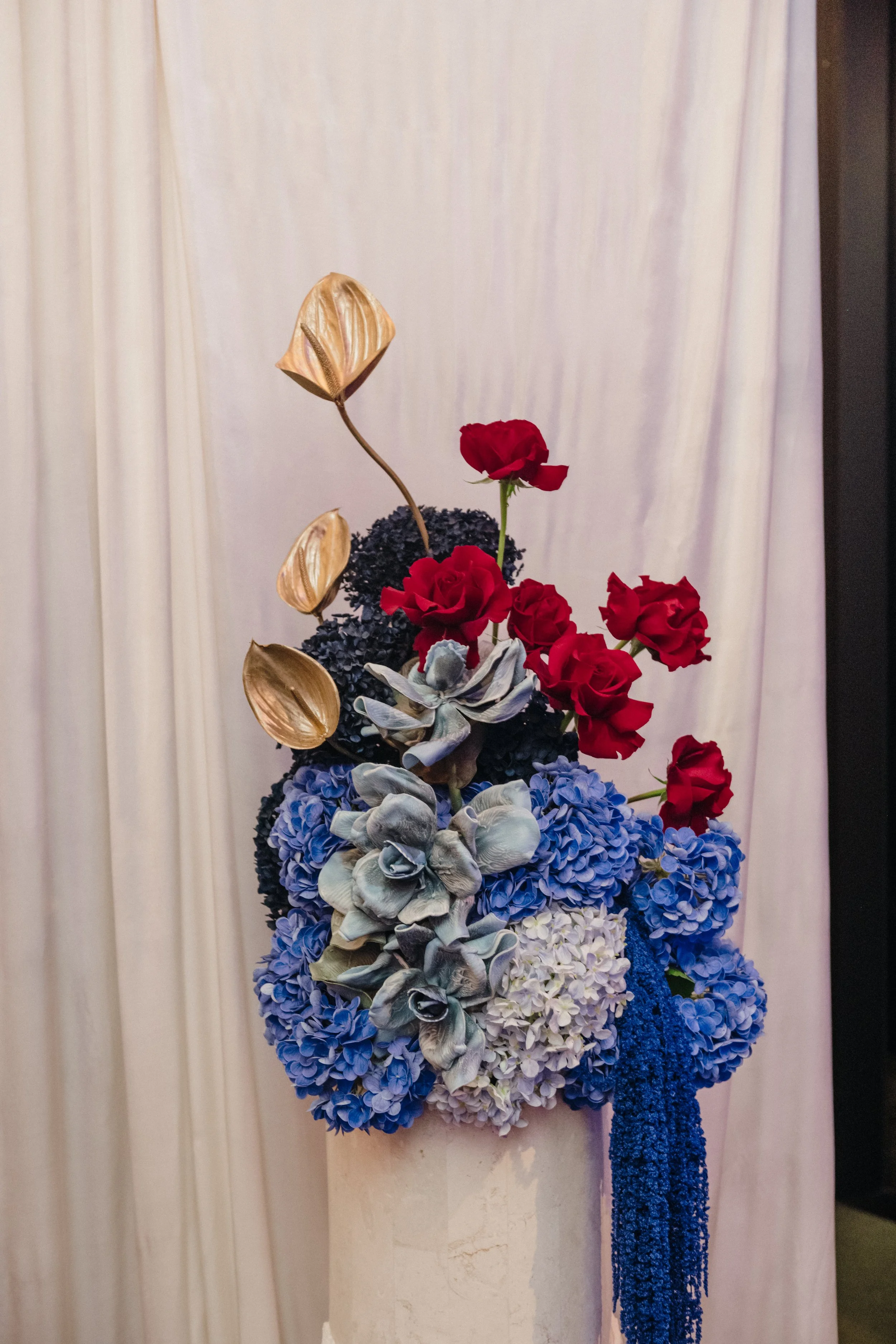 Modern‑lux corporate event styling by Poppy Culture at RACV Cape Schanck Resort, featuring a sculptural floral arrangement on a white pedestal against a soft draped backdrop. Vibrant red roses, metallic gold anthuriums, blue hydrangeas and blue‑grey 