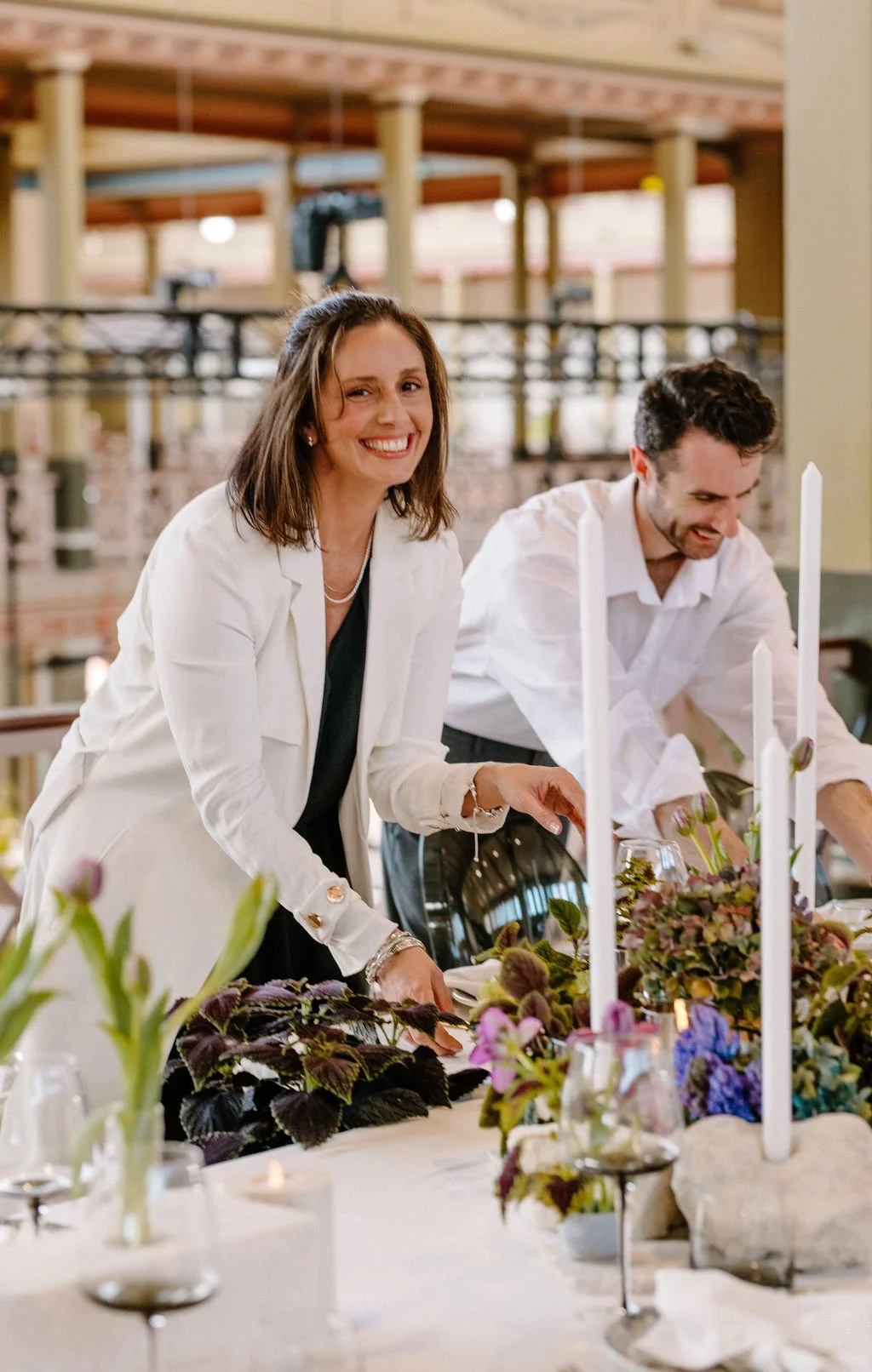 Alex & Lauren setting a formal table decorated with flowers and candles in a bright indoor space, smiling happily.