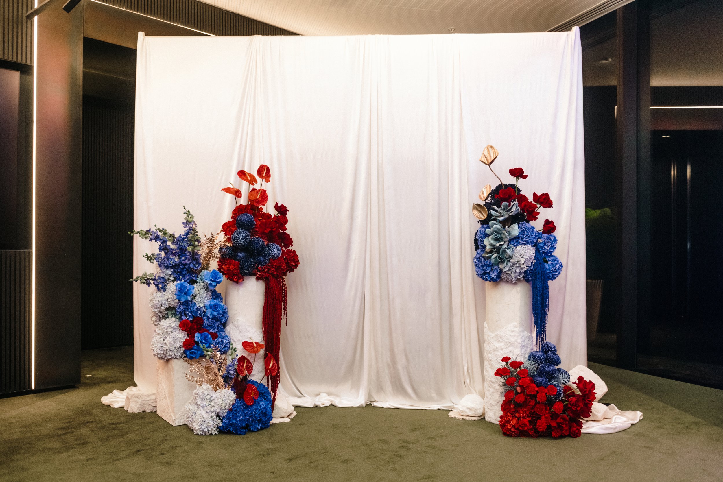 Modern‑lux corporate event styling by Poppy Culture at RACV Cape Schanck Resort, featuring two sculptural floral installations set against a white draped backdrop. Each cluster showcases blue hydrangeas, blue roses, red accent blooms, white florals a
