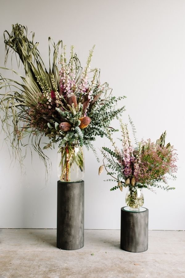Two large glass vases filled with pink, purple, and green floral arrangements on black cylindrical pedestals against a plain white wall.