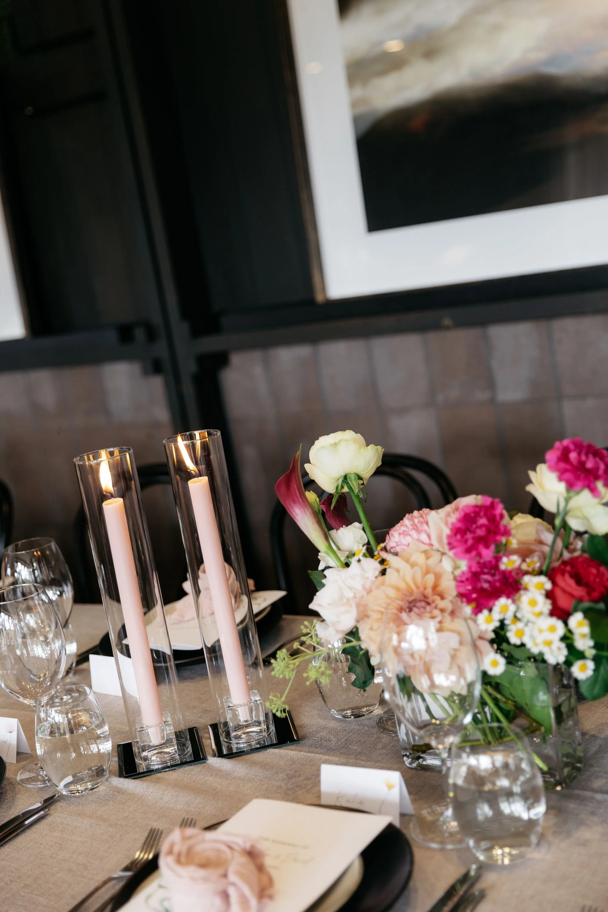 Intimate Polperro tablescape featuring sculptural pastel florals by Poppy Culture — including roses, dahlias, daisies and calla lilies — arranged alongside tall pink taper candles in glass holders, layered glassware, folded napkins and personalised n