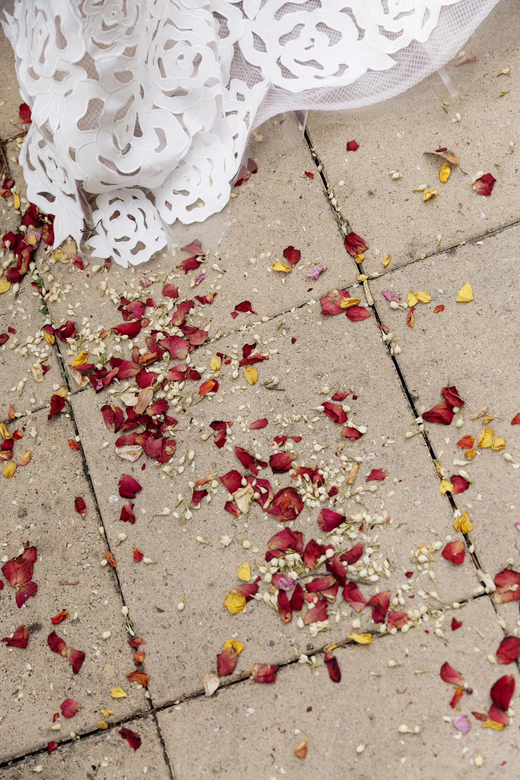 Close‑up wedding detail at Polperro capturing scattered flower petals and grains across textured tiles beneath the bride’s lace hem, with soft pastel tones and organic movement echoing the sculptural floral styling by Poppy Culture, beautifully docum