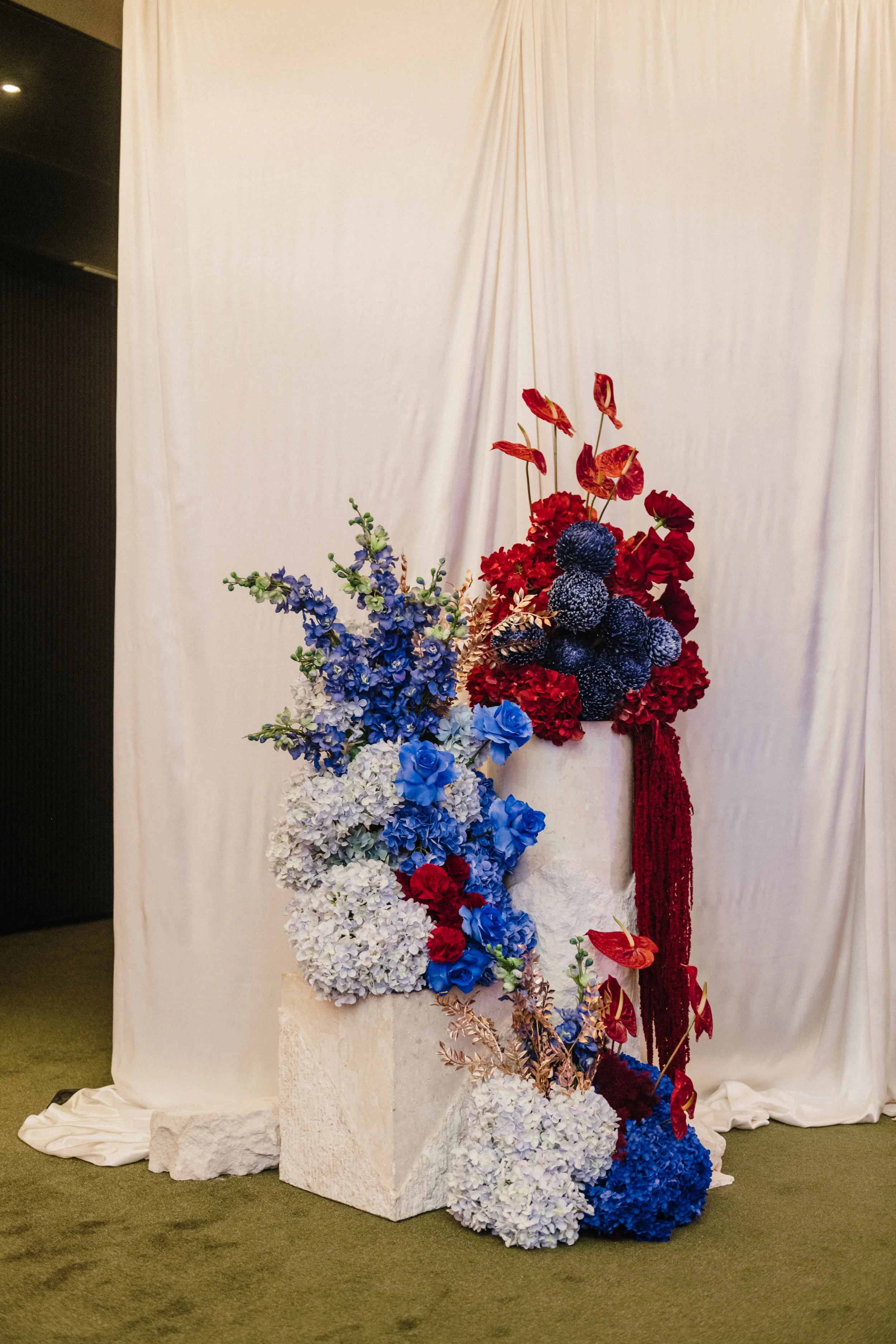 Modern‑lux corporate event styling by Poppy Culture at RACV Cape Schanck Resort, showcasing a vibrant floral installation against a white draped backdrop. Clusters of red anthuriums, blue hydrangeas, blue roses, white blooms and gold‑accent foliage a