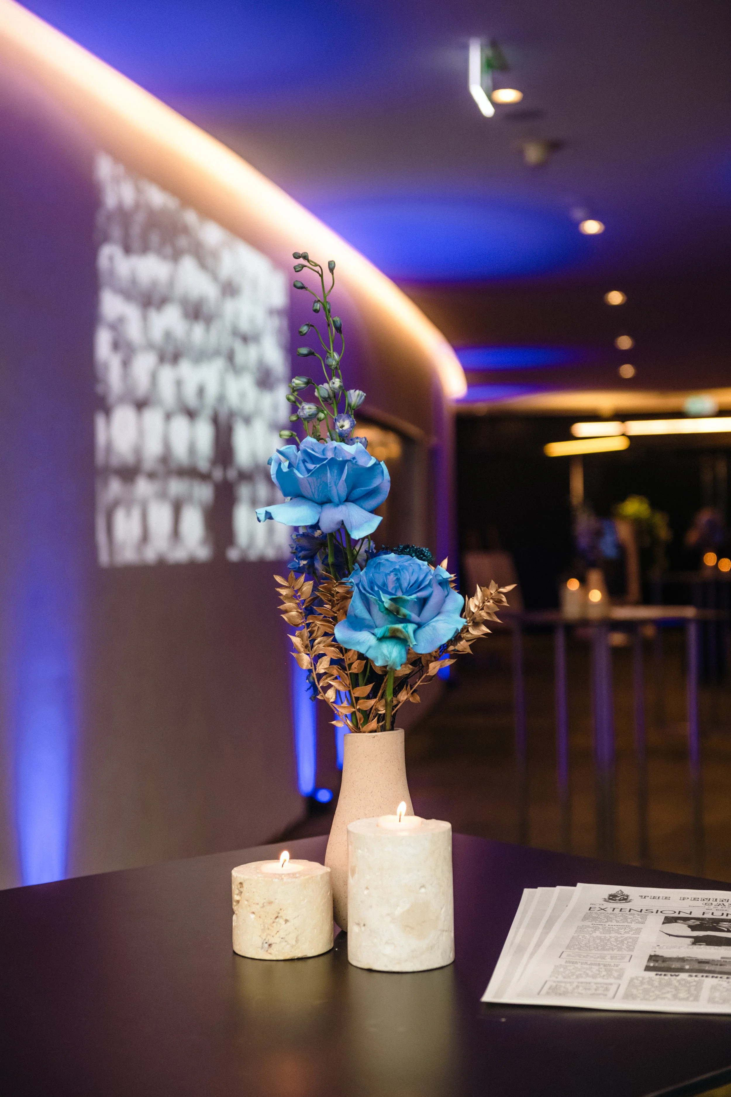 Modern‑lux corporate event styling by Poppy Culture at RACV Cape Schanck Resort, featuring a dark table vignette with a beige vase holding blue blooms and dried foliage. Two lit candles and a folded newspaper sit beside the arrangement, with a softly