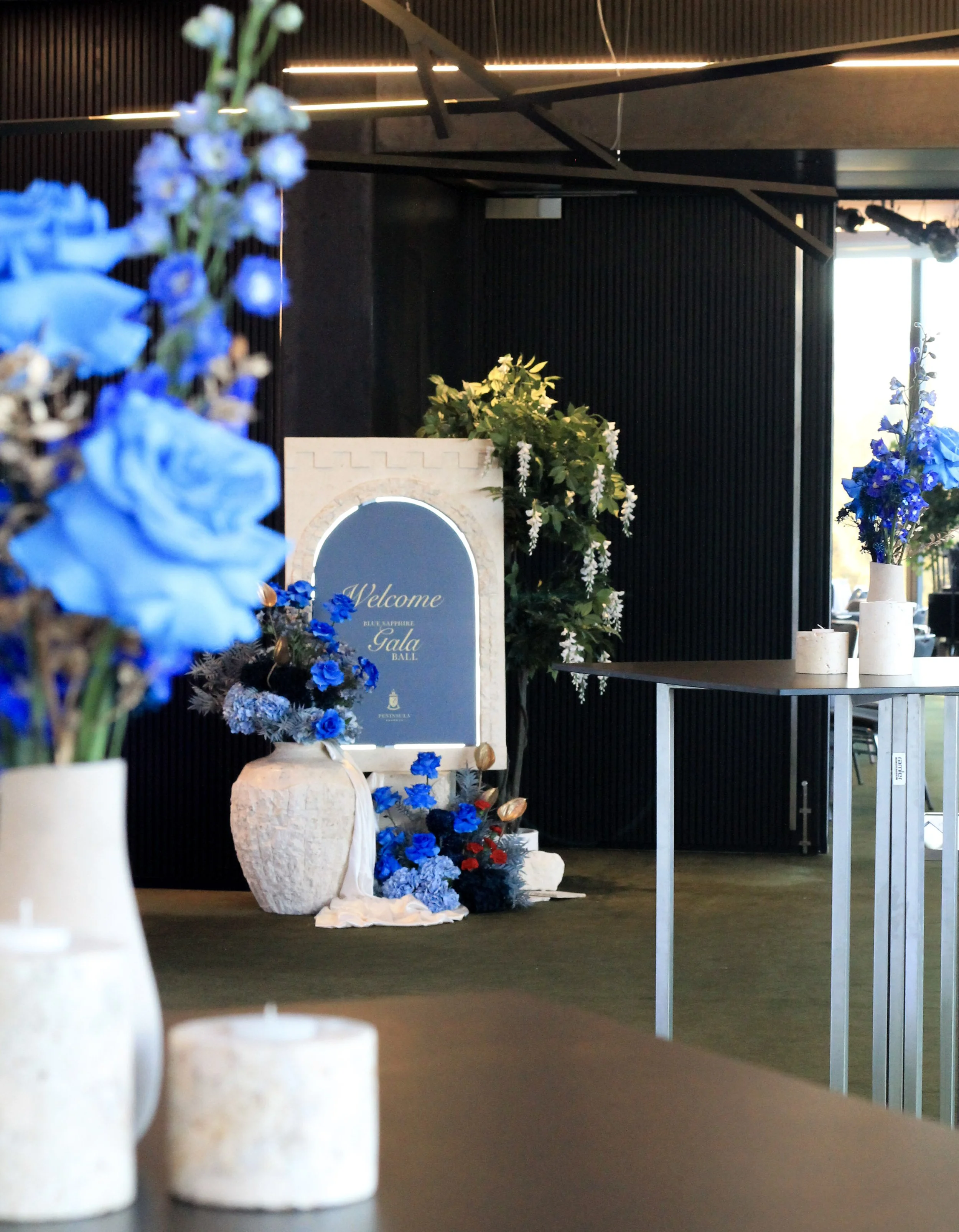 Modern‑lux corporate event styling by Poppy Culture at RACV Cape Schanck Resort, featuring blue and white floral arrangements with candles set on guest tables. In the background, a decorative “Welcome Gala” sign is framed by blue and white florals an