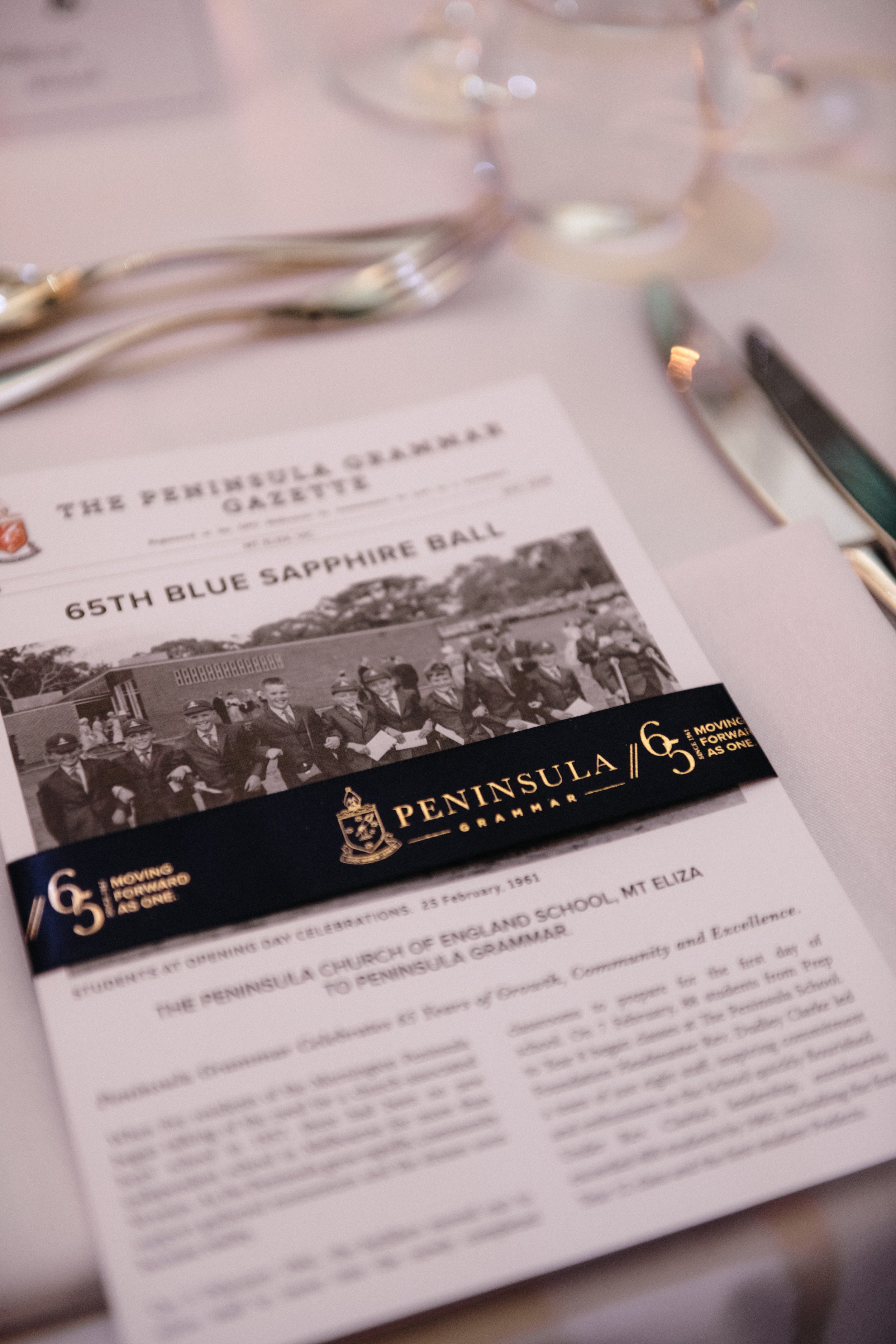 Modern‑lux corporate event styling by Poppy Culture at RACV Cape Schanck Resort, featuring a refined table setting with a Peninsula Grammar Gazette program placed on a white napkin. The design highlights the “65th Blue Sapphire Ball” with a deep‑blue