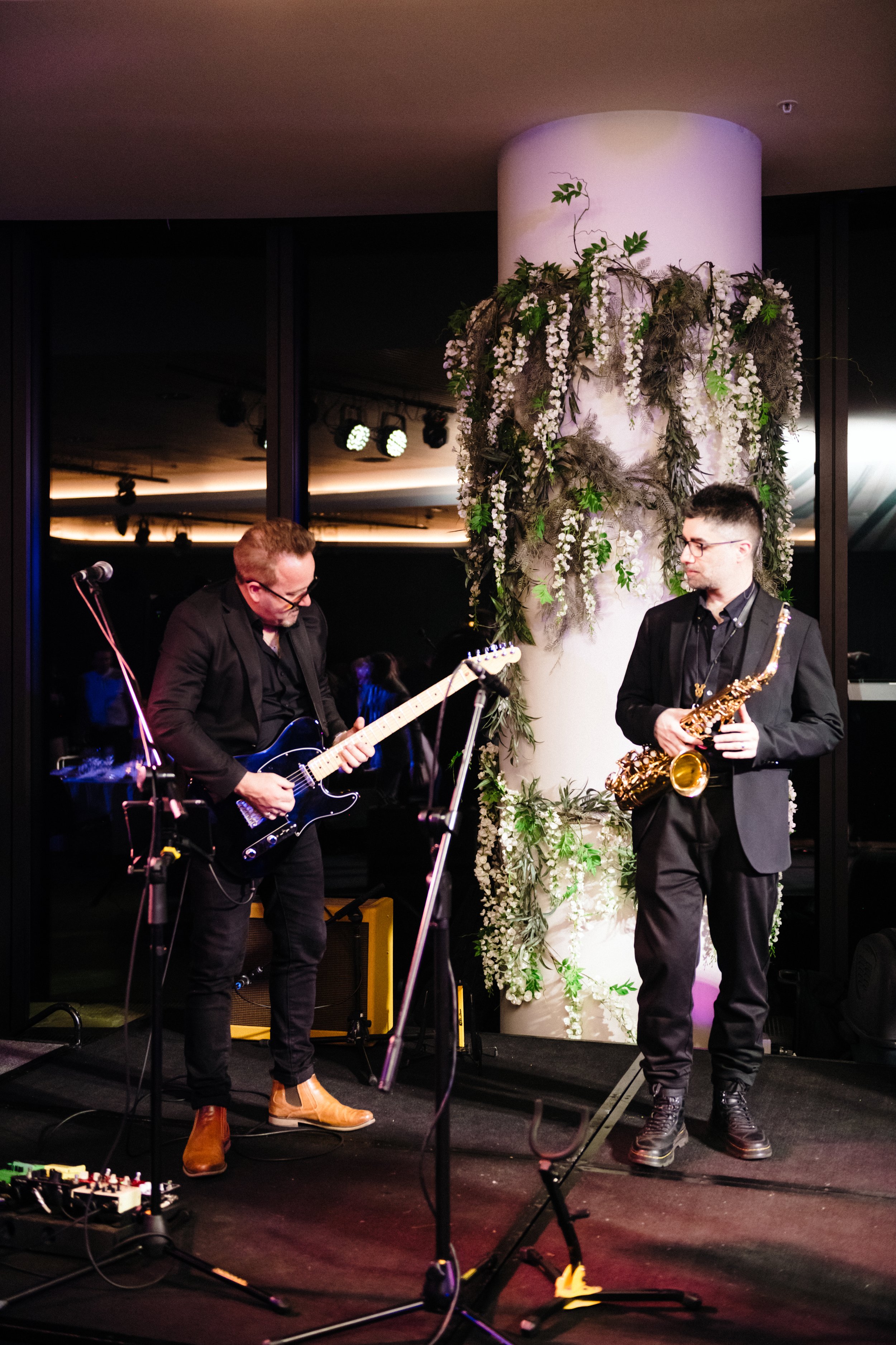 Modern‑lux corporate event styling by Poppy Culture at RACV Cape Schanck Resort, featuring a live performance by the Baker Boys Band. Musicians on drums, keyboard and bass perform on a softly lit stage framed by greenery and reflective windows. The e