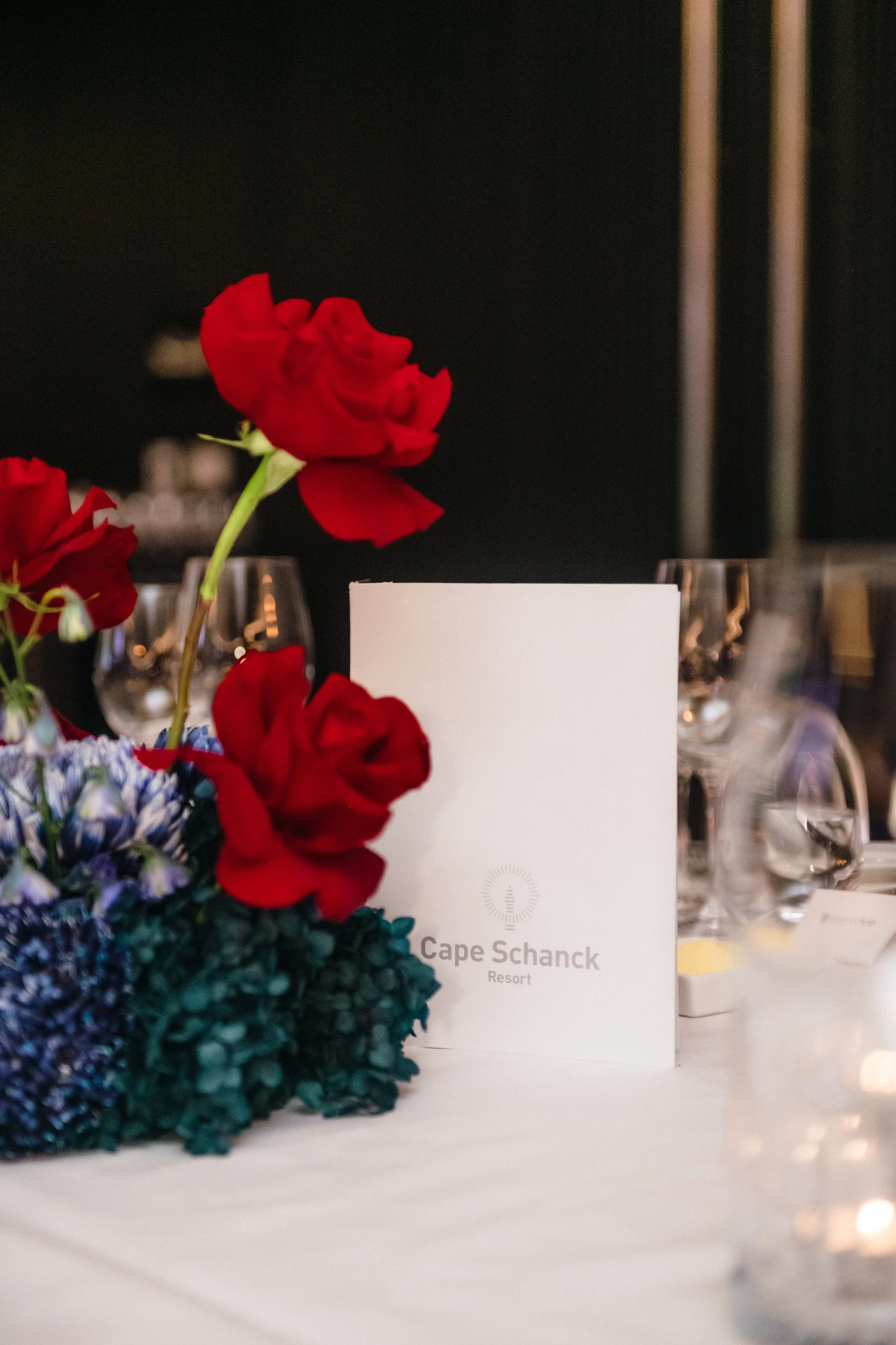 Modern‑lux corporate event styling by Poppy Culture at RACV Cape Schanck Resort, featuring an elegant table vignette with a Cape Schanck Resort menu card framed by red roses and deep‑blue hydrangeas. Soft candlelight and refined glassware create a wa