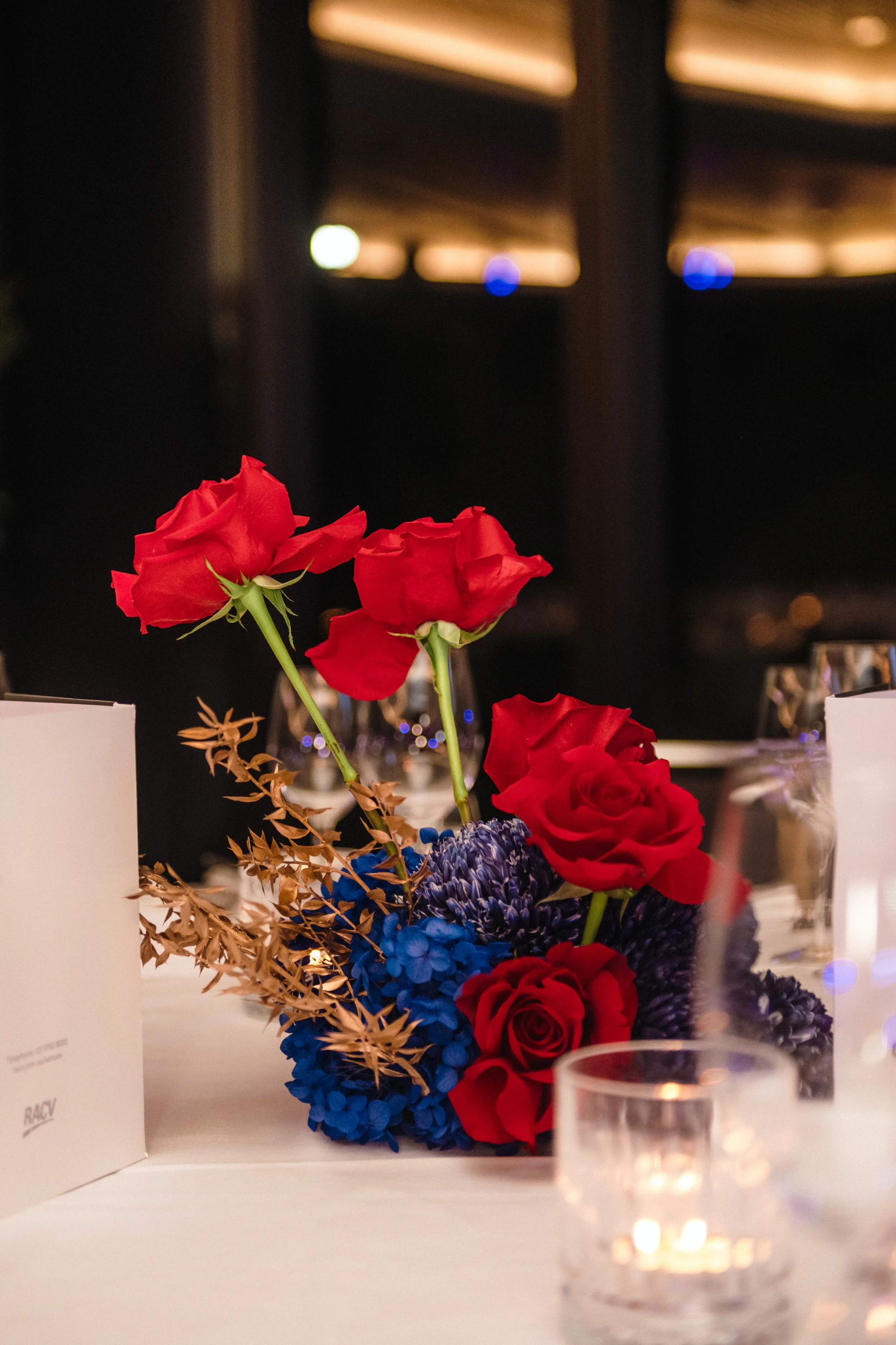 Modern‑lux corporate event styling by Poppy Culture at RACV Cape Schanck Resort, featuring a compact table arrangement of red roses, deep‑blue blooms and metallic gold foliage. Soft candlelight, refined glassware and folded menu cards sit on crisp wh
