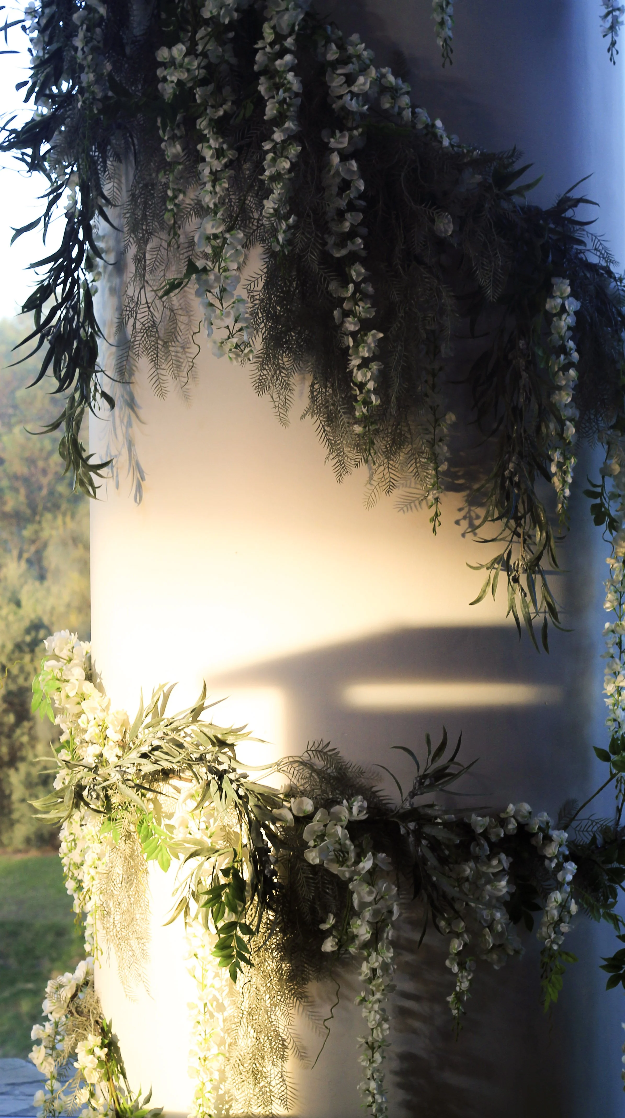  
Modern‑lux corporate event styling by Poppy Culture at RACV Cape Schanck Resort, featuring a white cylindrical column adorned with cascading greenery and delicate white florals. Warm sunlight creates soft shadows across the textured foliage, with 
