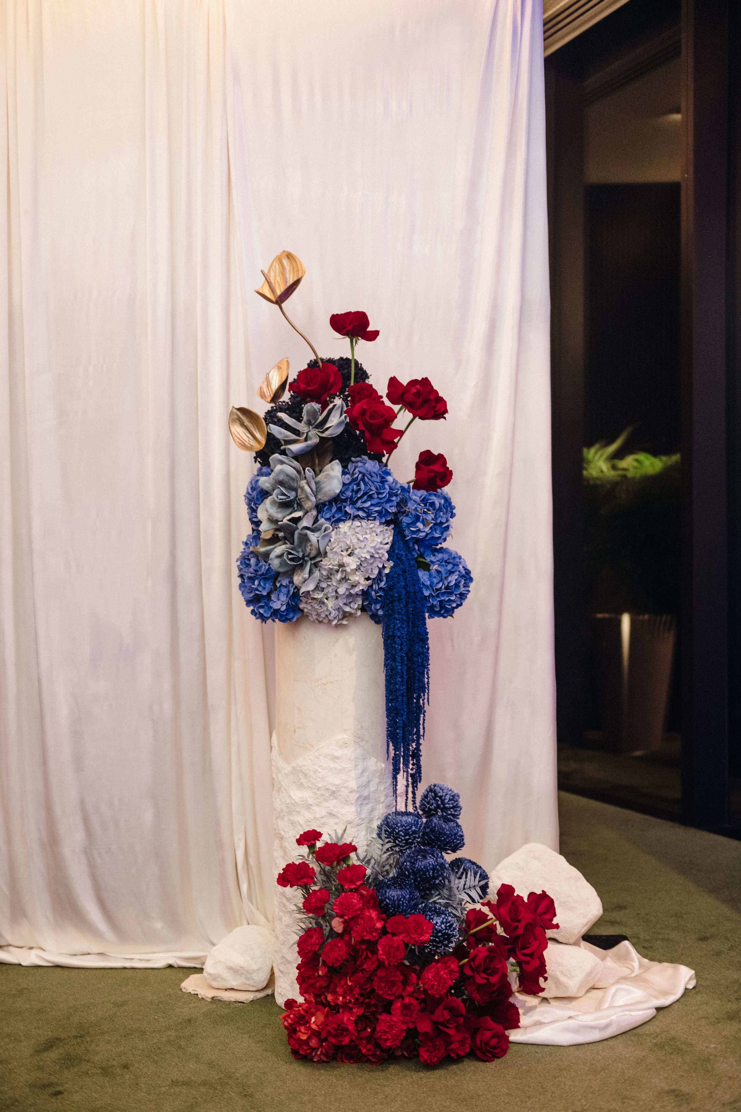 Modern‑lux corporate event styling by Poppy Culture at RACV Cape Schanck Resort, featuring a tall sculptural floral installation on a white draped pedestal. Red roses, blue hydrangeas, metallic gold blooms and cascading deep‑blue detailing create hei