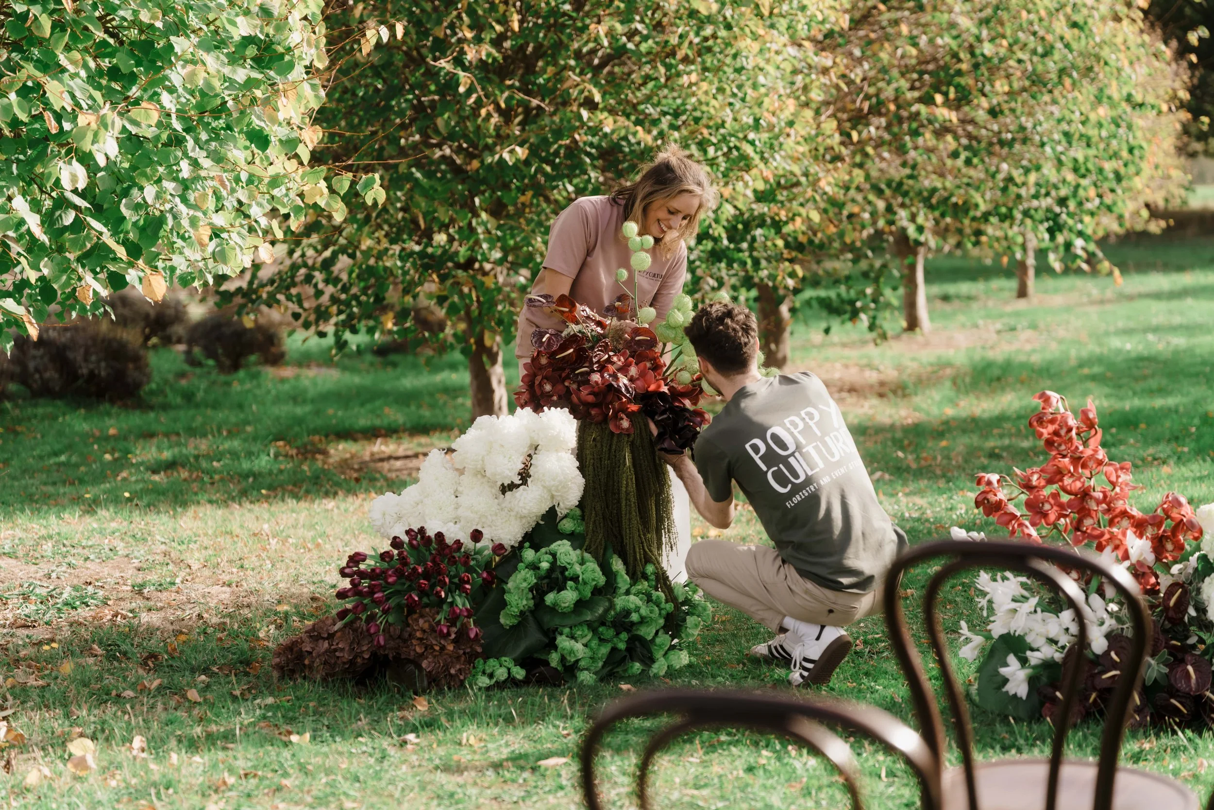 Alex and Lauren working on a flower arrangement outdoors. They are surrounded by various colourful flowers and trees in the background.