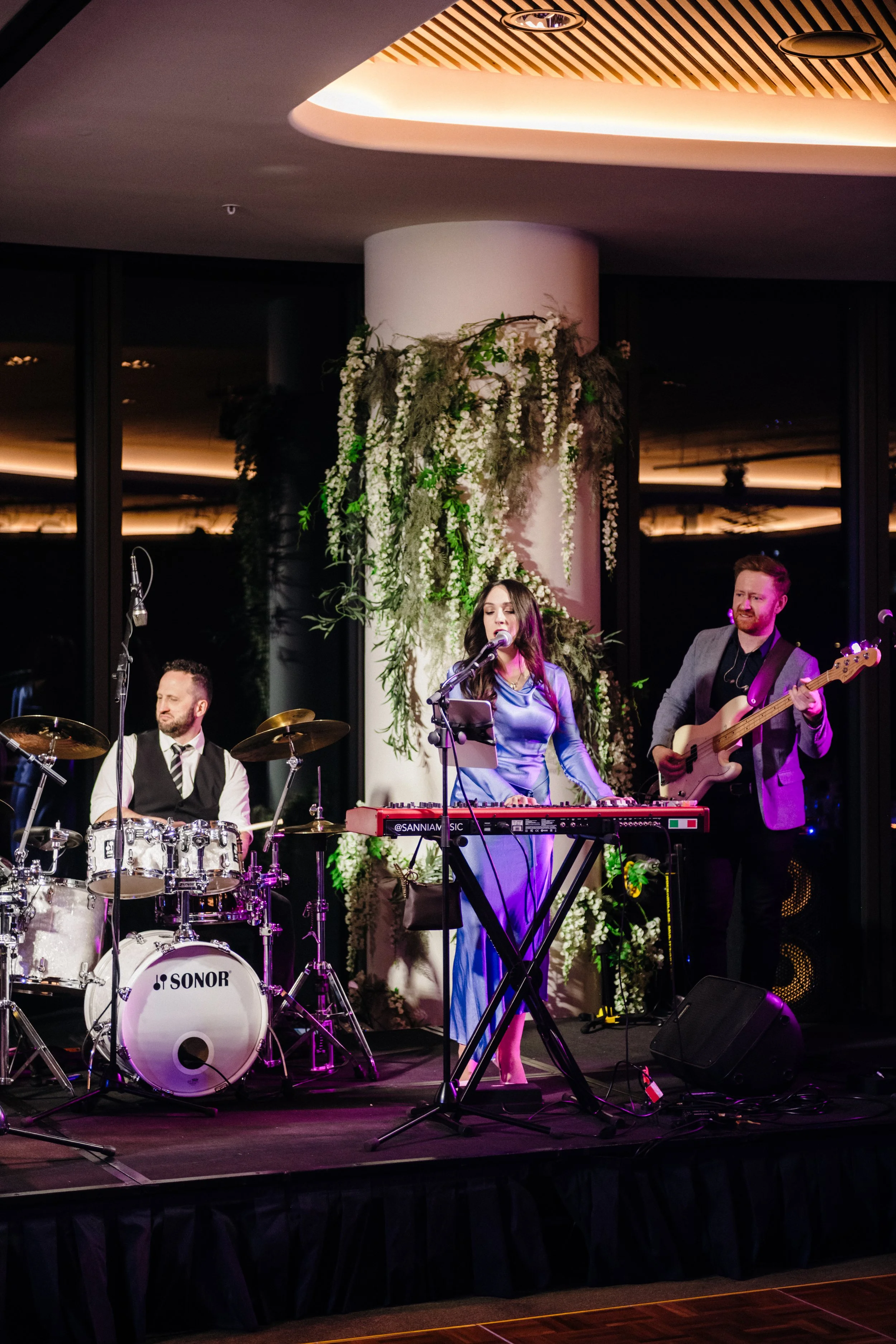 Modern‑lux corporate event styling by Poppy Culture at RACV Cape Schanck Resort, featuring a live performance by the Baker Boys Band. Musicians on drums, keyboard and bass perform on a softly lit stage framed by greenery and reflective windows. The e