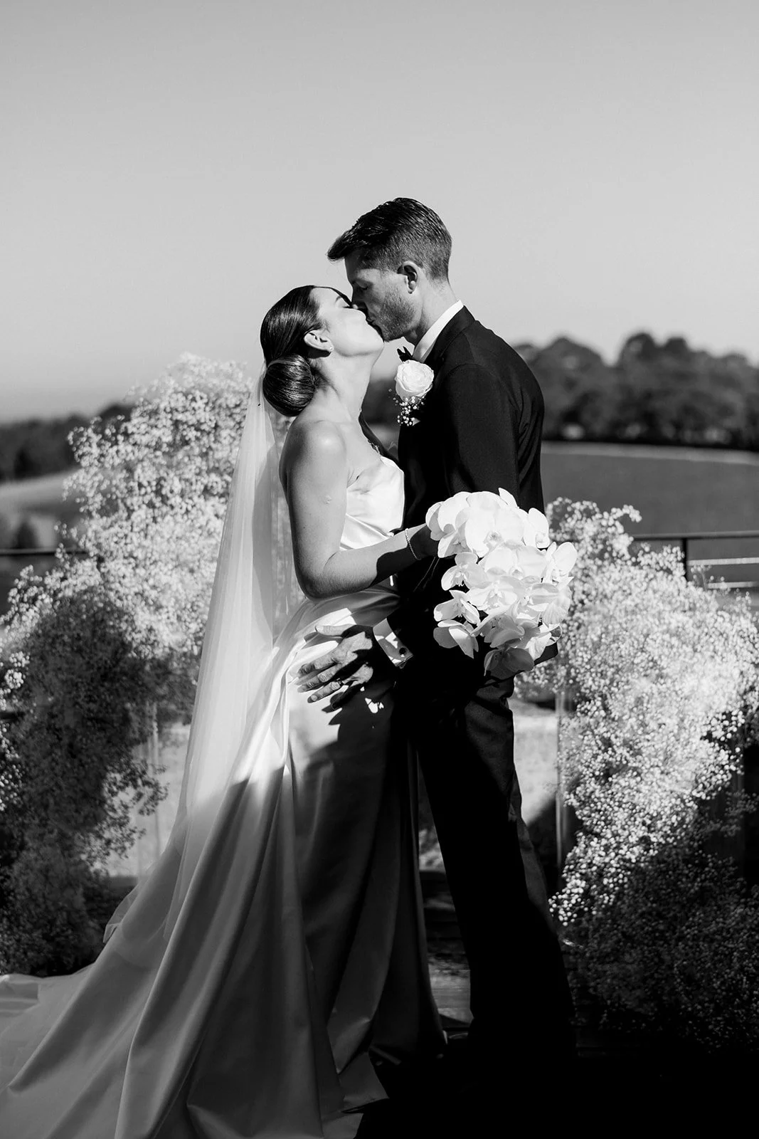 Black‑and‑white modern editorial wedding moment at Port Phillip Estate, with the couple sharing a kiss against the vineyard landscape and white florals styled by Poppy Culture.