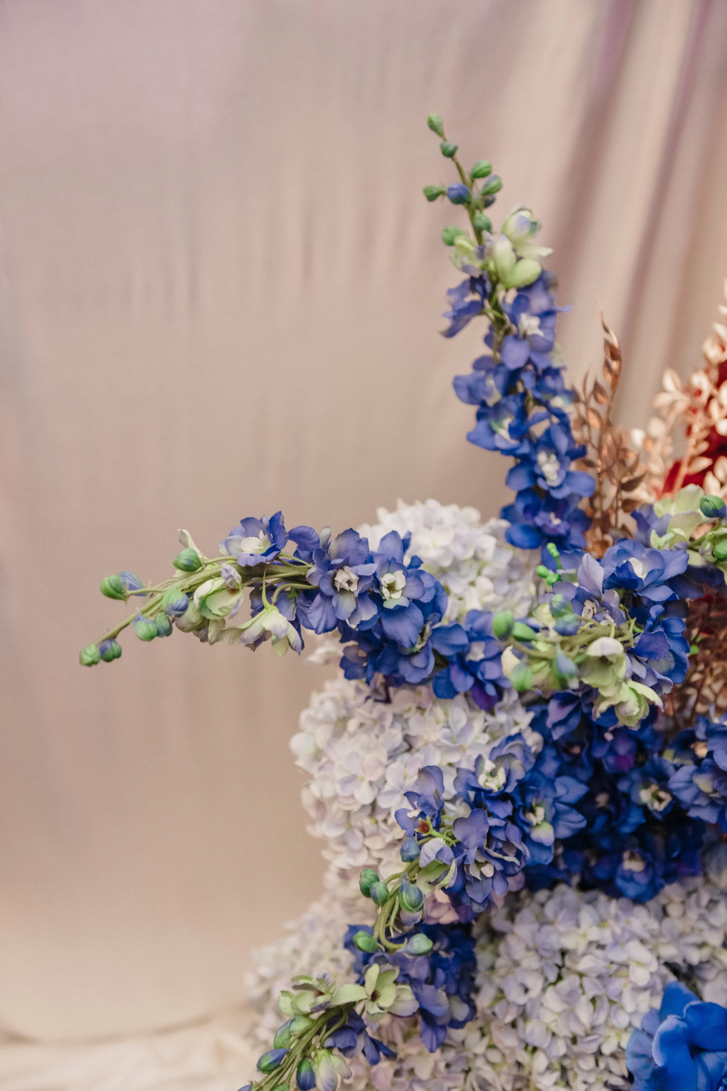 Modern‑lux corporate event styling by Poppy Culture at RACV Cape Schanck Resort, featuring a soft beige backdrop with clustered blue and lilac blooms. Hydrangeas and delphinium‑style florals are layered with dried brown foliage, creating a refined co