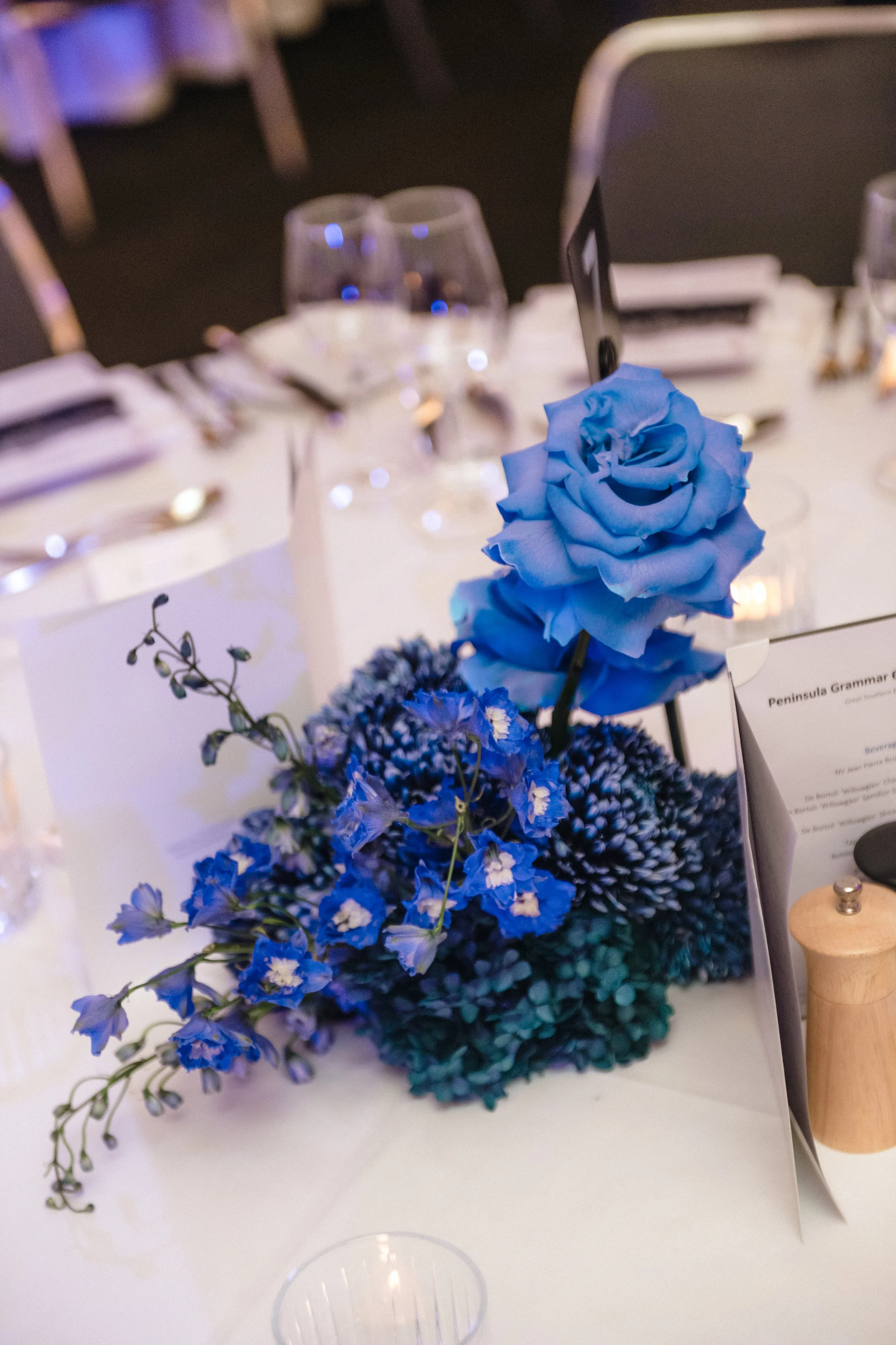 Modern‑lux corporate event styling by Poppy Culture at RACV Cape Schanck Resort, featuring a compact blue‑toned table centerpiece with a vivid blue rose surrounded by smaller textured blooms. Refined glassware, folded napkins, menu cards and a wooden