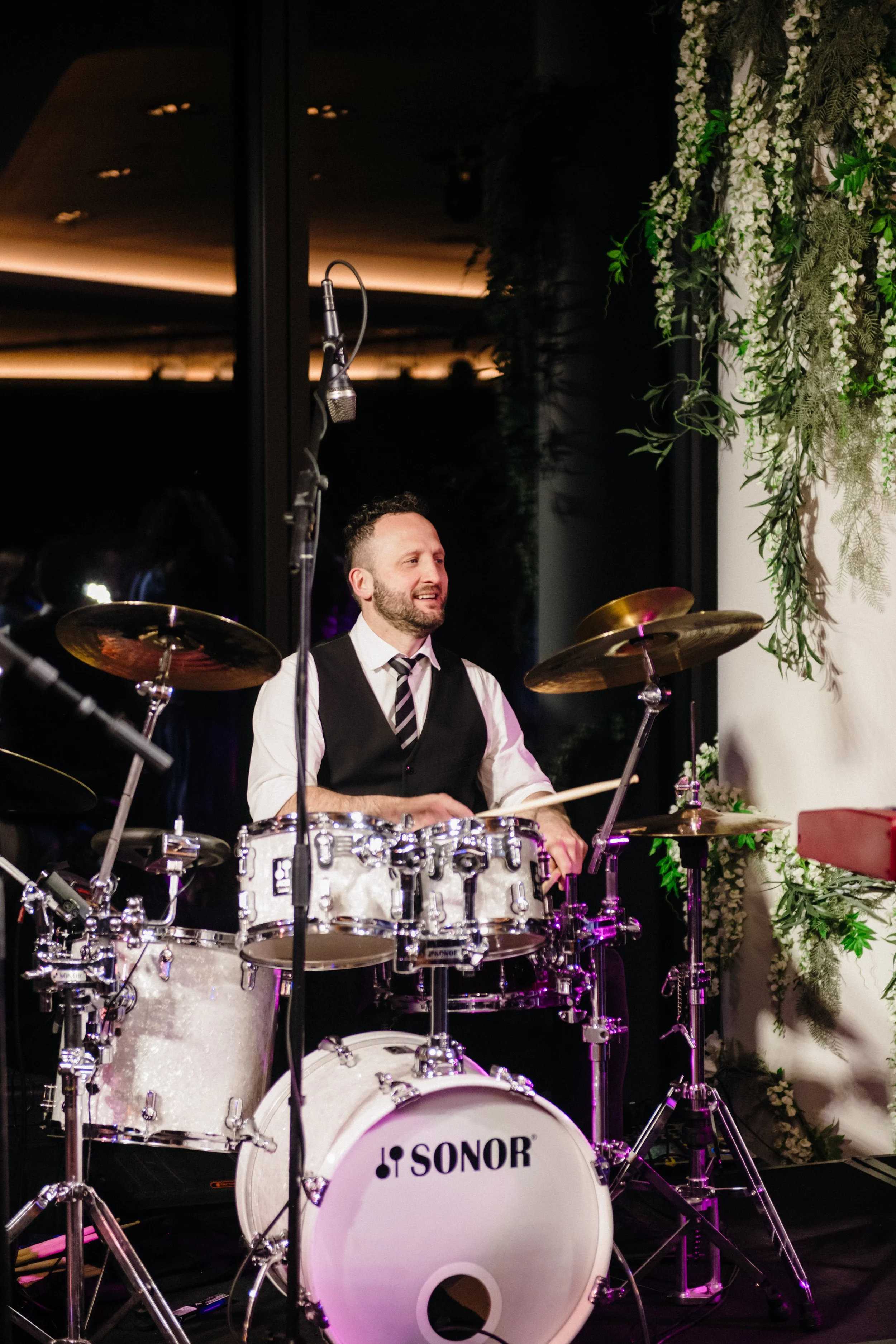 Modern‑lux corporate event styling by Poppy Culture at RACV Cape Schanck Resort, featuring a live performance by the Baker Boys Band. Musicians on drums, keyboard and bass perform on a softly lit stage framed by greenery and reflective windows. The e