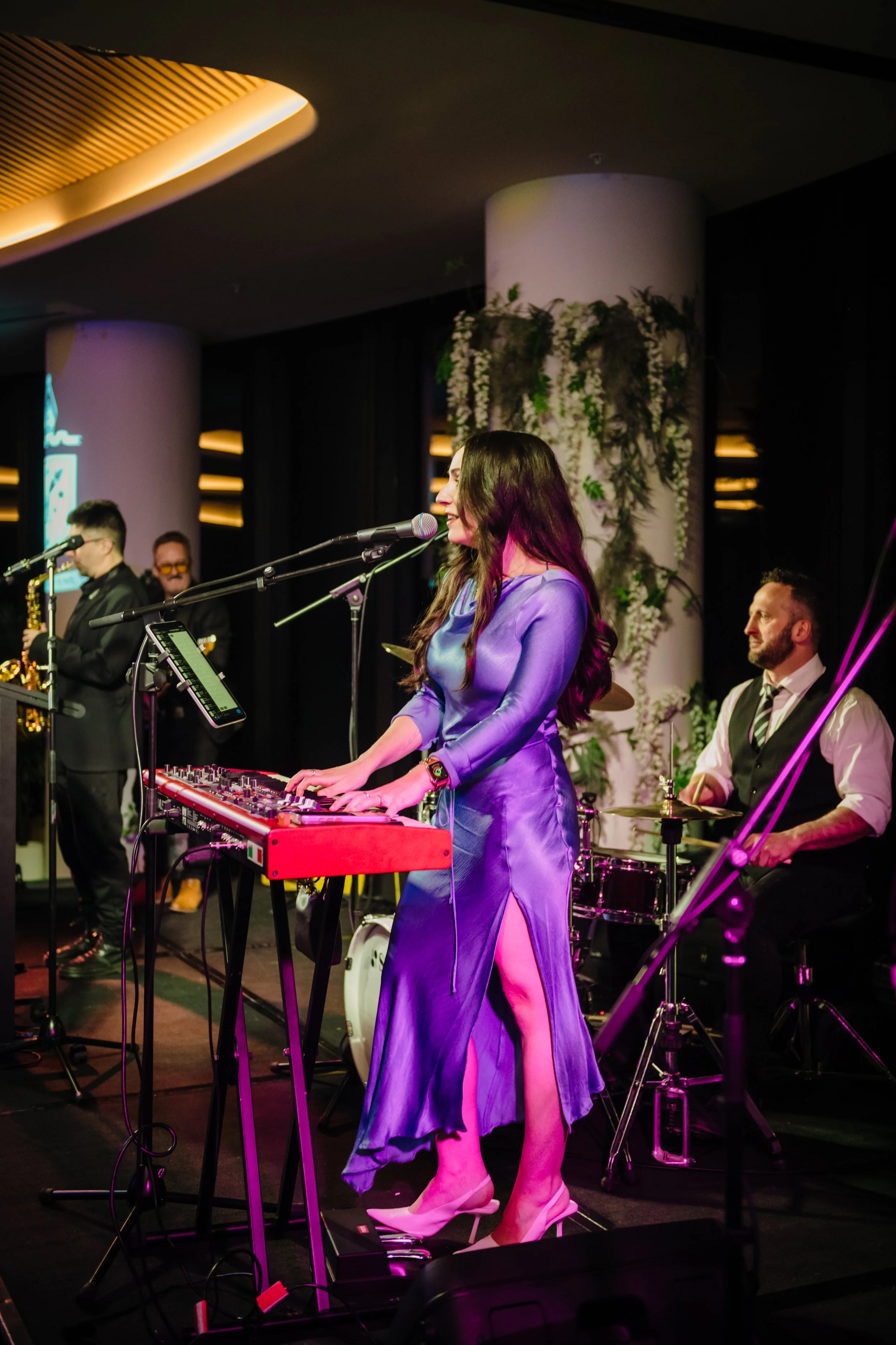 Modern‑lux corporate event styling by Poppy Culture at RACV Cape Schanck Resort, featuring a live performance by the Baker Boys Band. Musicians on drums, keyboard and bass perform on a softly lit stage framed by greenery and reflective windows. The e