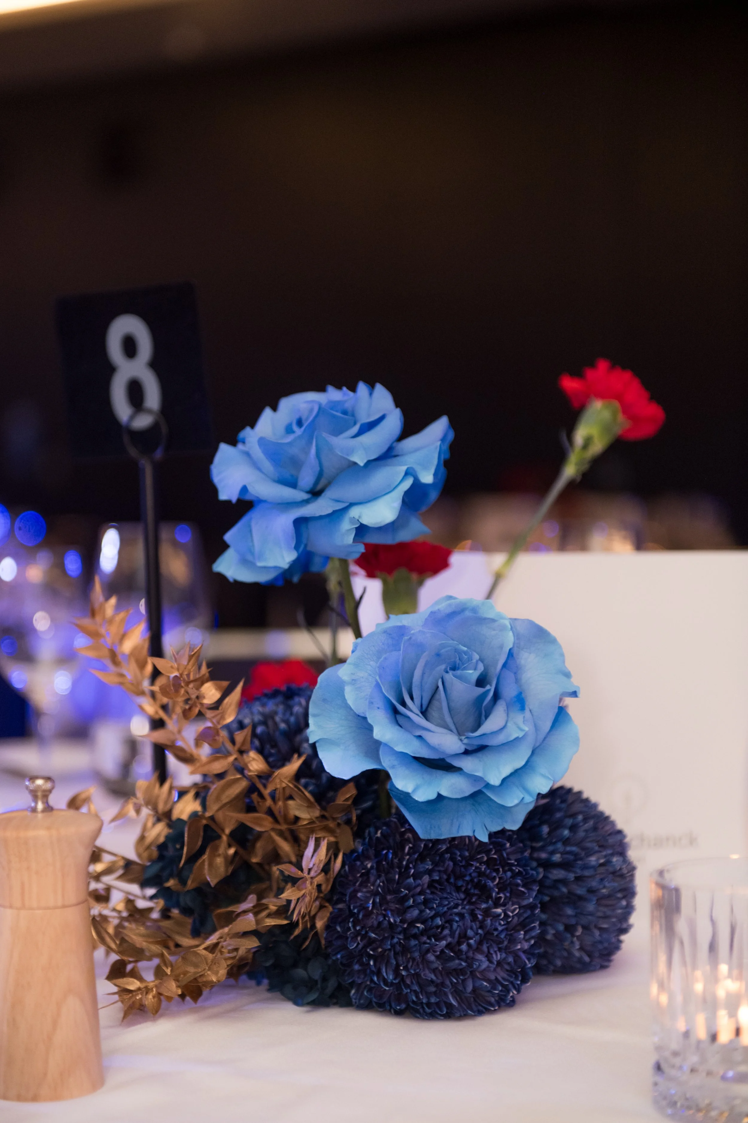 Modern‑lux corporate event styling by Poppy Culture at RACV Cape Schanck Resort, featuring a bold table centerpiece with two vivid blue roses, red accent blooms and metallic gold foliage arranged in a deep‑blue base. Refined glassware, a white place 