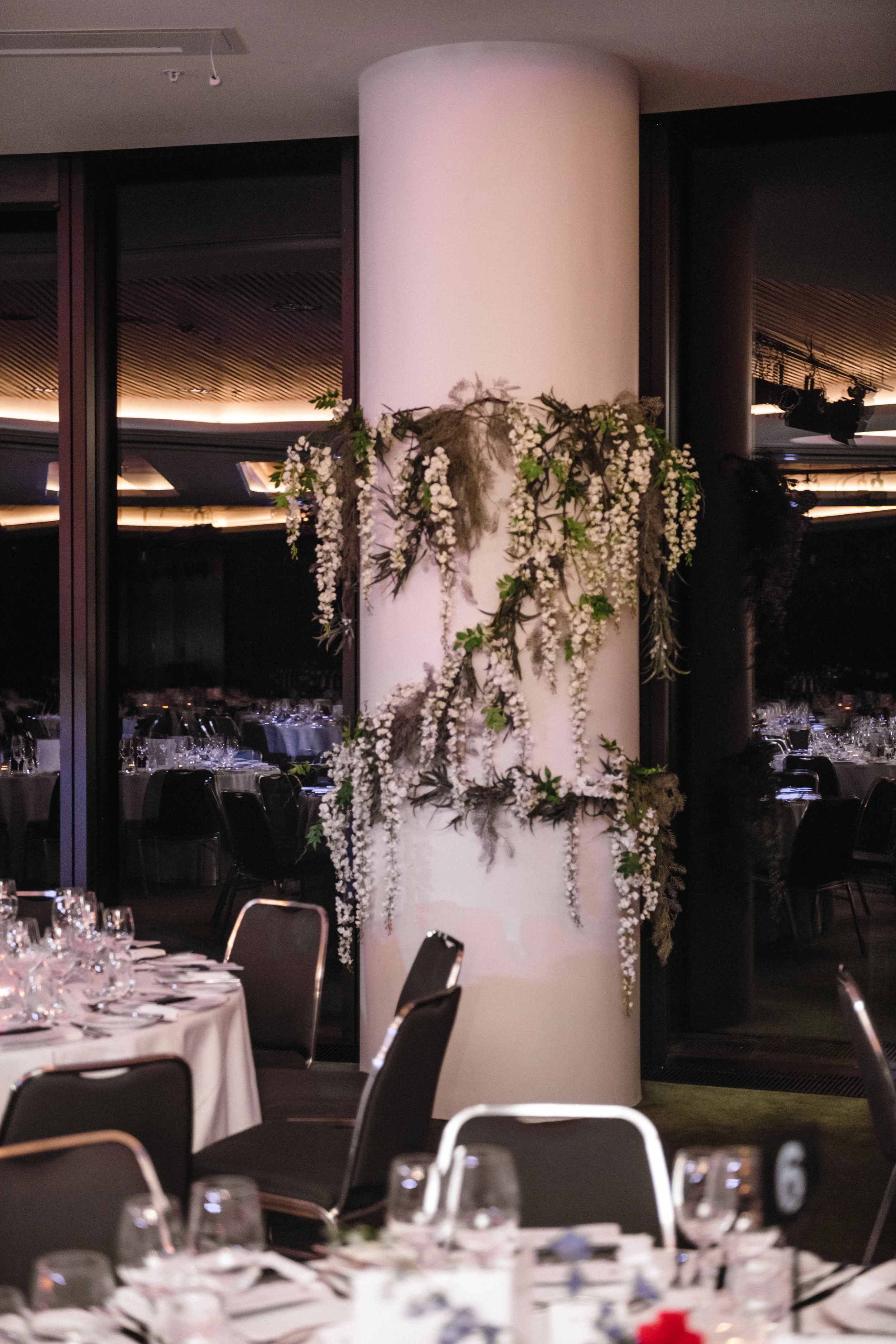 Modern‑lux corporate event styling by Poppy Culture at RACV Cape Schanck Resort, featuring an elegant dining space with round tables dressed in white linens and refined glassware. A central white cylindrical column is adorned with cascading greenery 