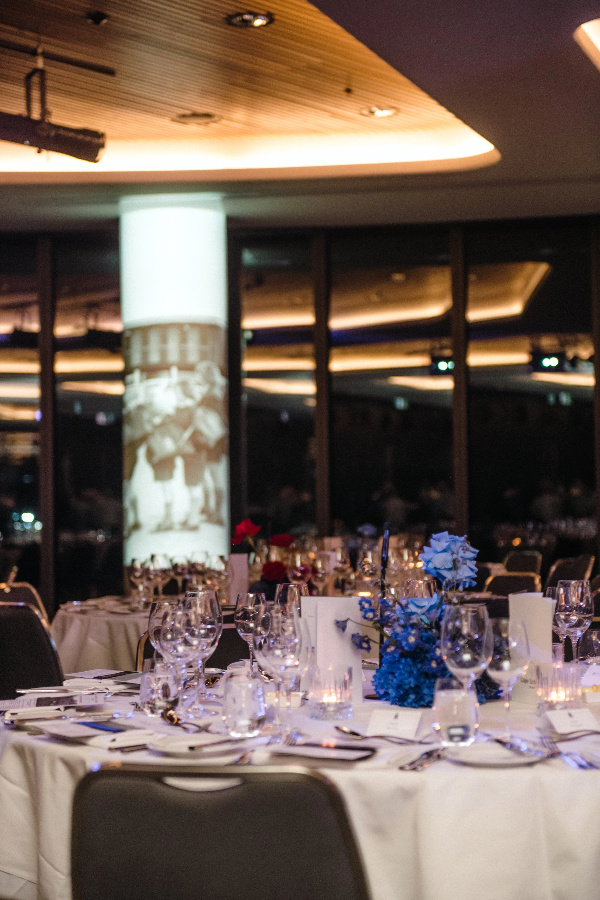   
Modern‑lux corporate event styling by Poppy Culture at RACV Cape Schanck Resort, featuring a formal dining space with round tables dressed in white linens, layered glassware and refined place settings. Blue floral centrepieces add colour to the ro