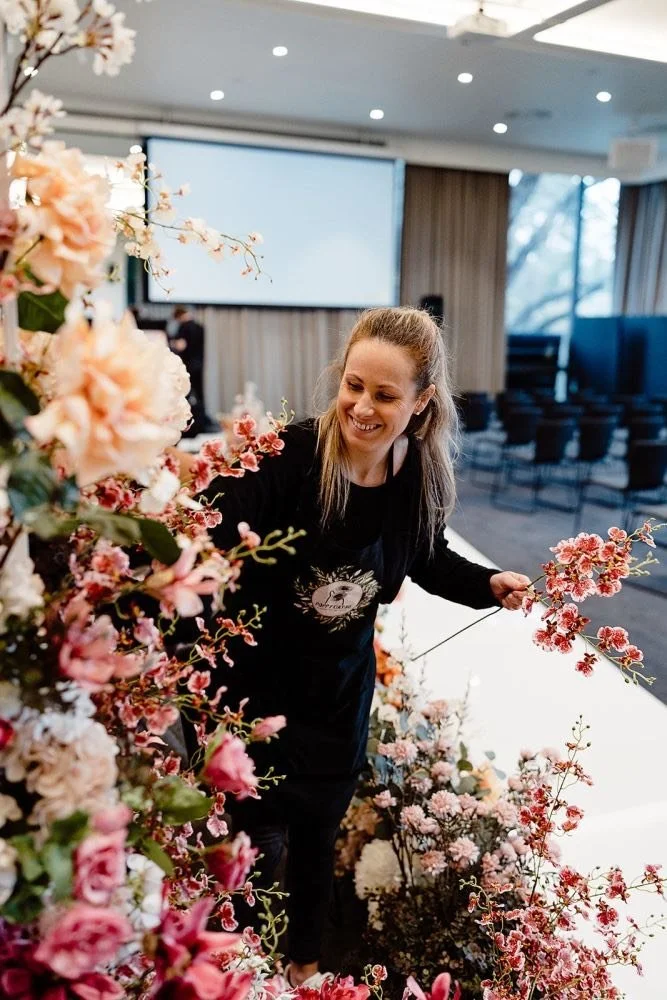Lauren arranging pink and white flowers on a stage at an indoor event venue.