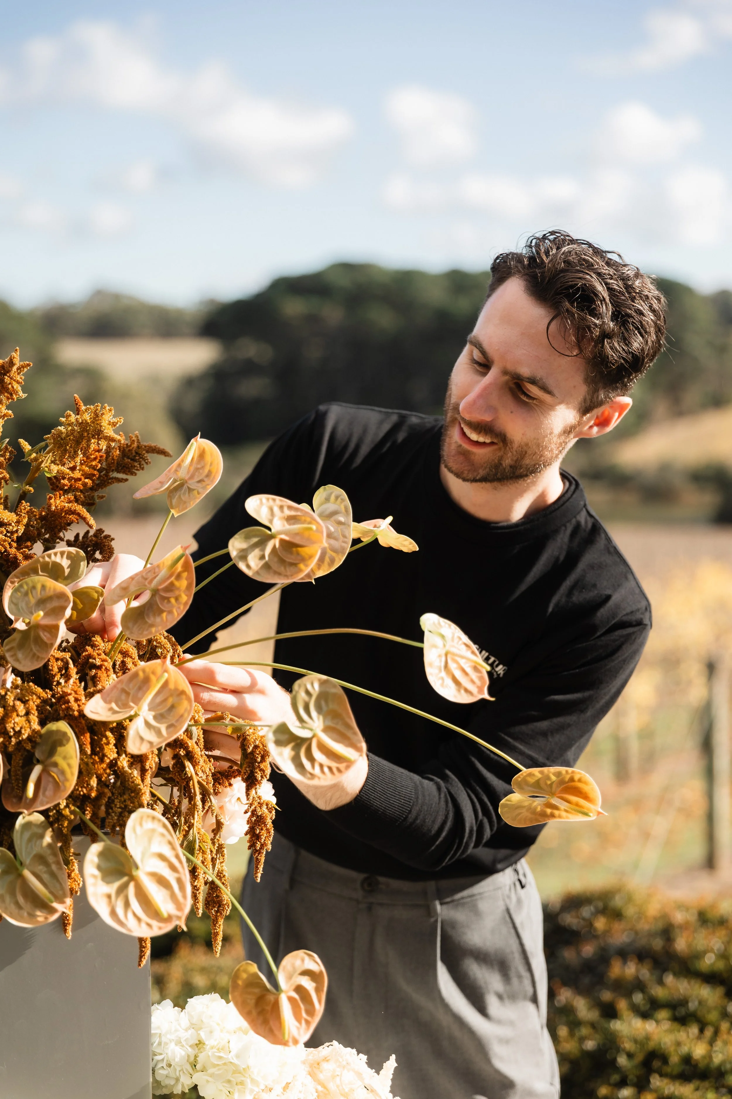 Alex is arranging large, orange, and yellow flowers outdoors on a sunny day.