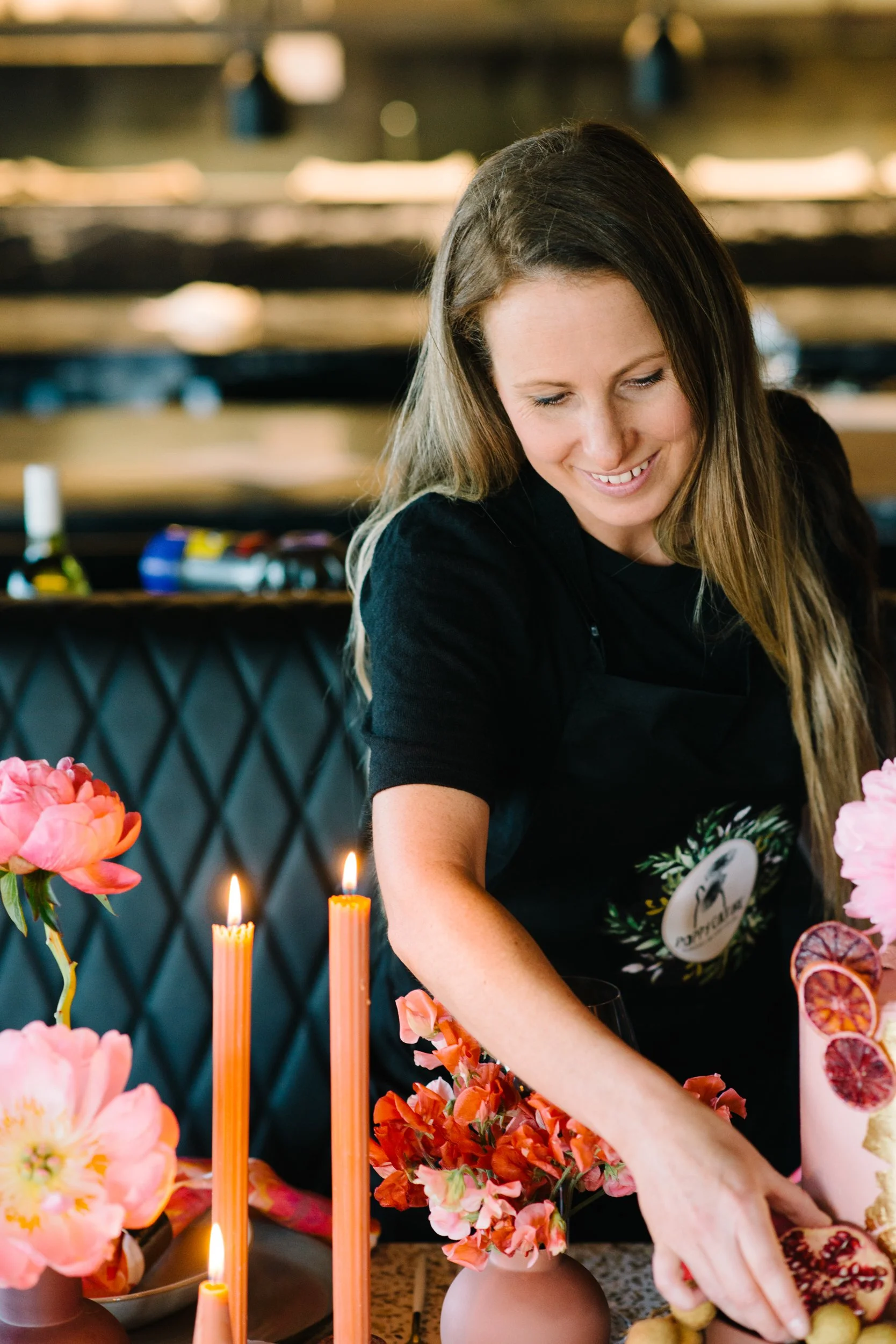 Lairen arranging flowers on a table decorated with pink peonies, orange flowers, pink candles, and sliced citrus fruits in a tall pink drink.