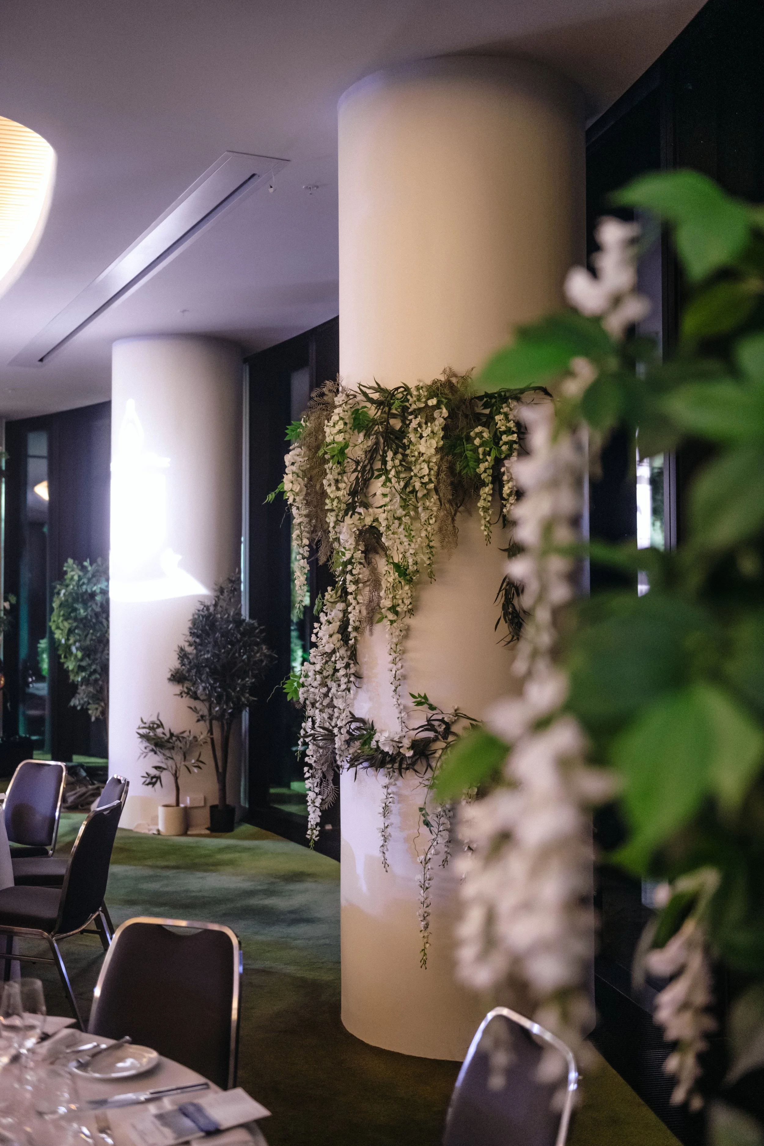 Modern‑lux corporate event styling by Poppy Culture at RACV Cape Schanck Resort, featuring an indoor dining space with white cylindrical columns dressed in cascading greenery and white florals. Tables are set with refined place settings, glassware an
