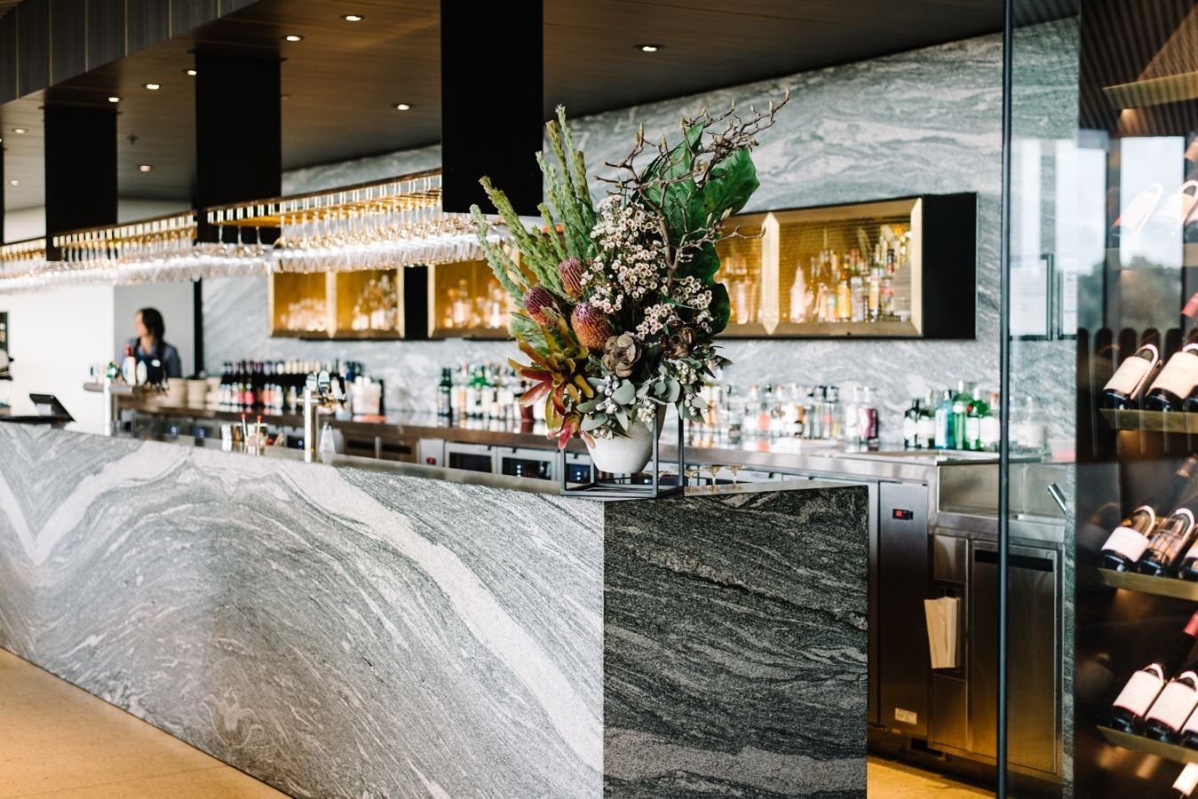 Modern bar with a marble counter, a large floral arrangement in a white vase, and a bartender in the background. Glass shelves with liquor bottles and wine racks are also visible.