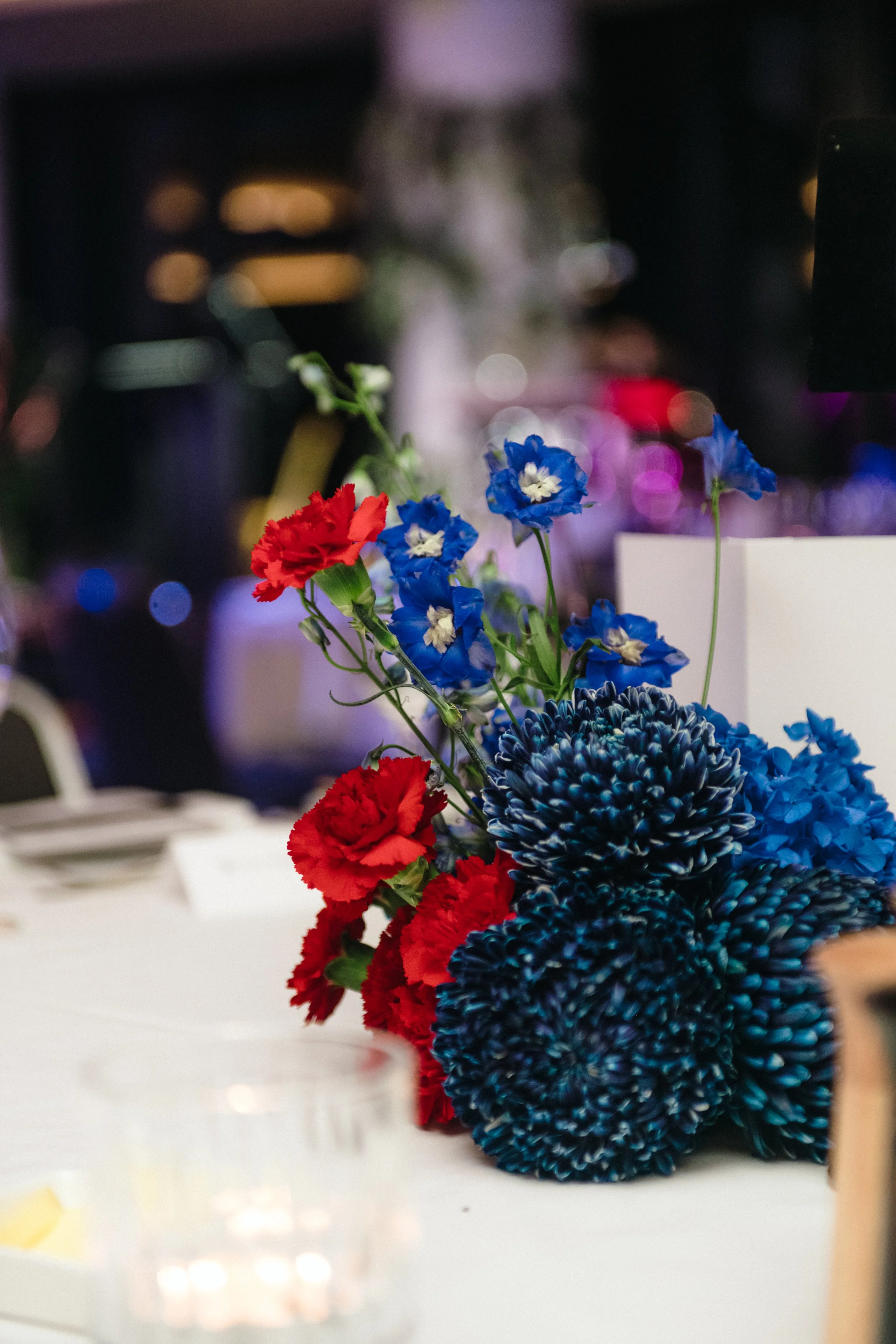 Modern‑lux corporate event styling by Poppy Culture at RACV Cape Schanck Resort, featuring a vibrant table arrangement of red roses, blue chrysanthemums and complementary textured blooms. The blurred ambient lighting and surrounding tables create an 