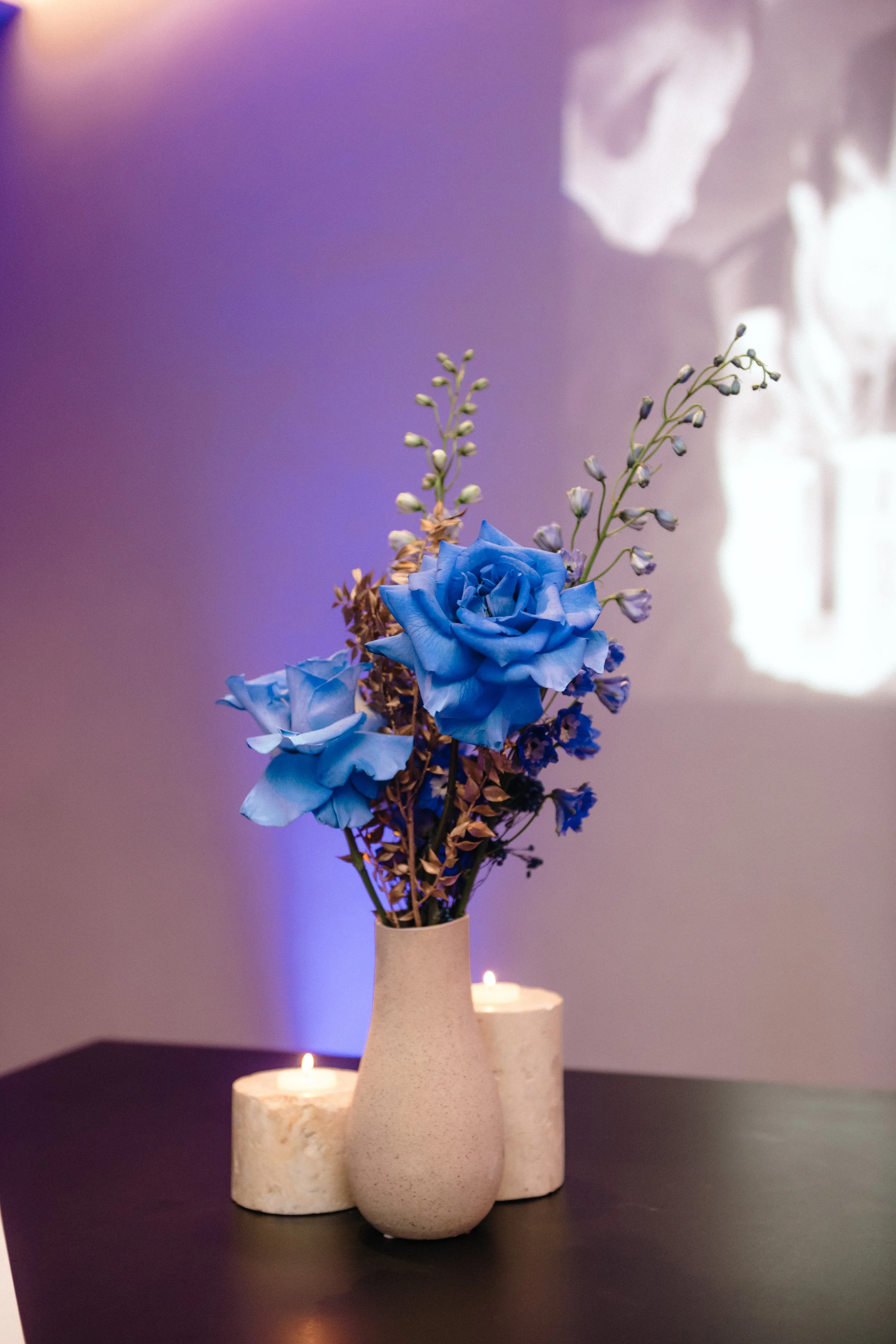 Modern‑lux corporate event styling by Poppy Culture at RACV Cape Schanck Resort, featuring a beige vase arrangement of vivid blue roses with soft white and purple blooms. Two lit candles sit behind the vignette on a dark surface, with a softly lit pu
