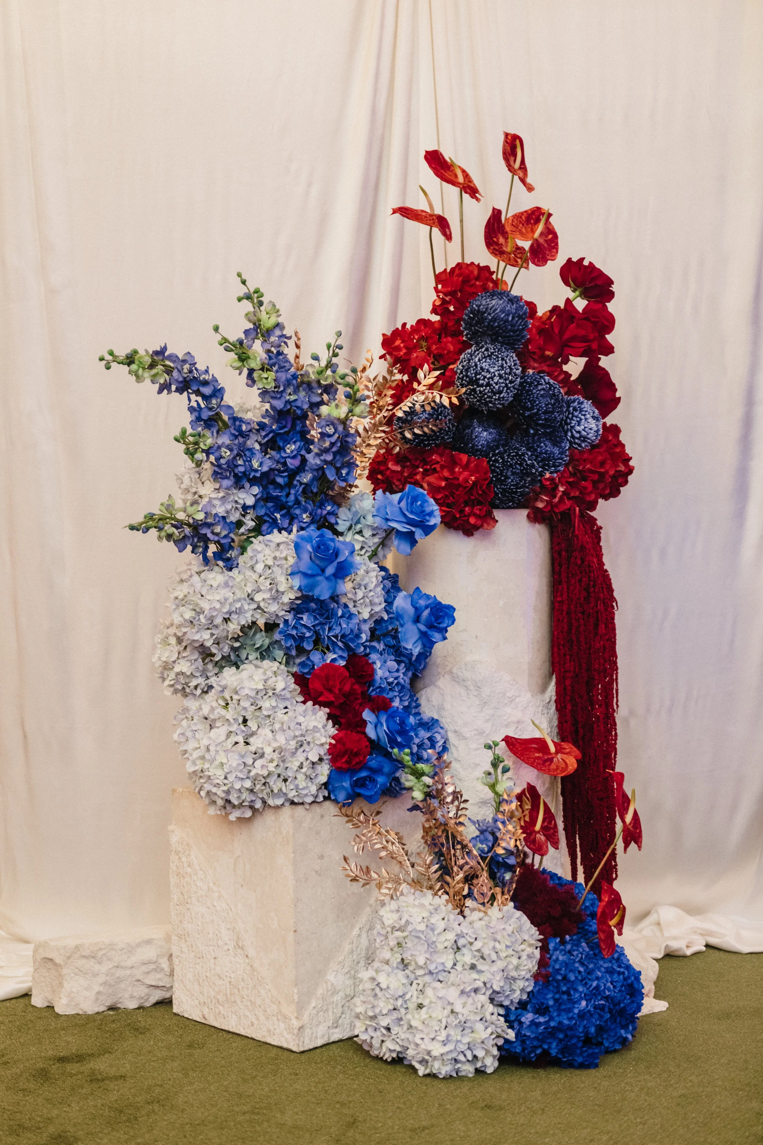 Modern‑lux corporate event styling by Poppy Culture at RACV Cape Schanck Resort, featuring a tiered floral installation set against a white draped backdrop. Clusters of red anthuriums, blue hydrangeas, blue roses, white blooms and textured delphinium