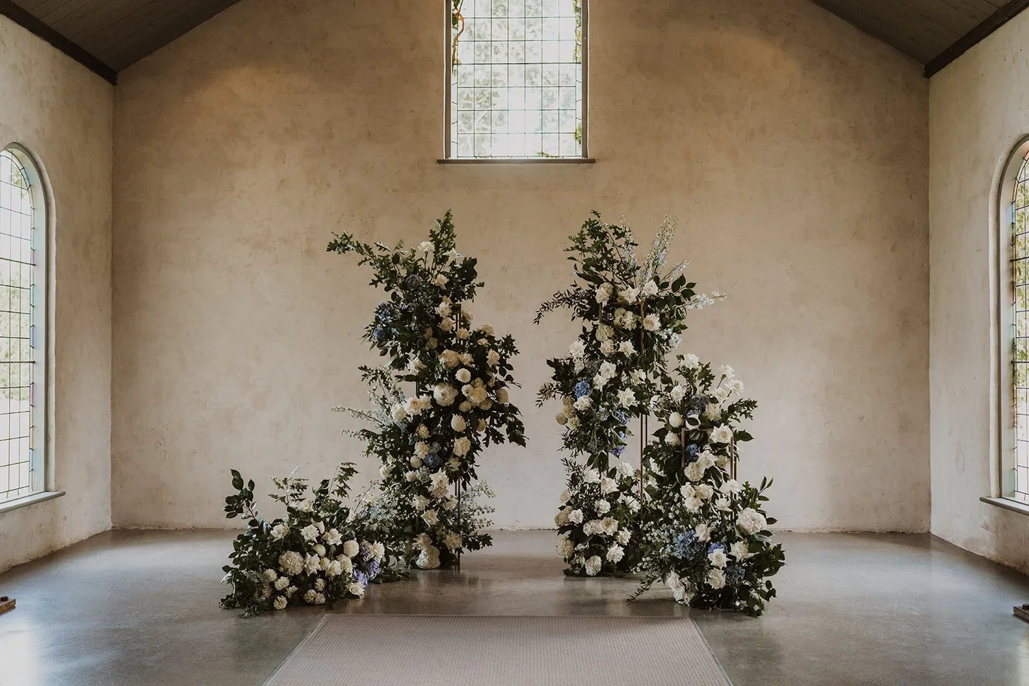 Poppy Culture wedding florals at Stones of the Yarra Valley with white and blue arrangements beneath arched stained‑glass windows.