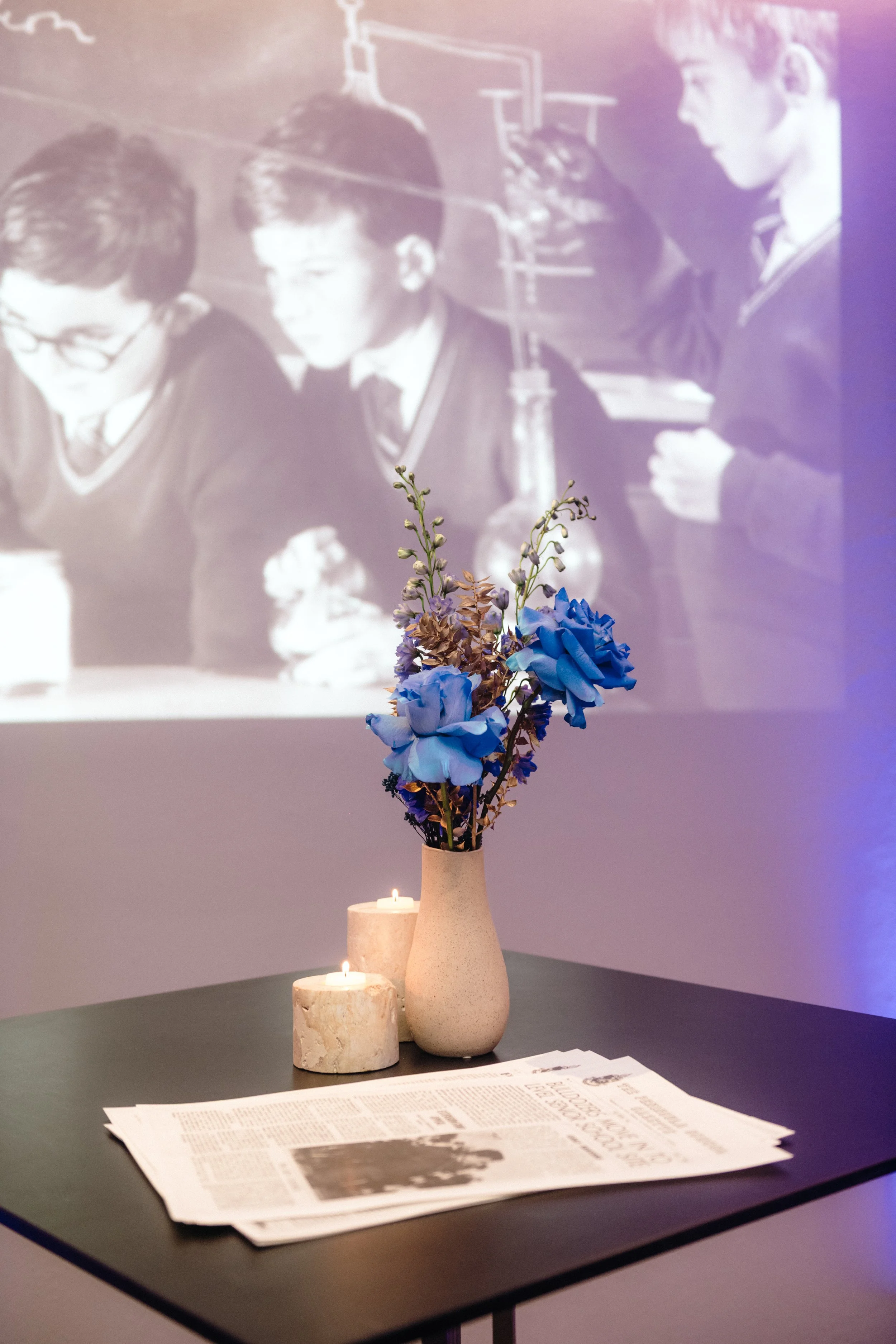 Modern‑lux corporate event styling by Poppy Culture at RACV Cape Schanck Resort, featuring a tabletop vignette with a beige vase of blue and purple blooms, two lit candles and a folded newspaper on a dark surface. In the background, a black‑and‑white