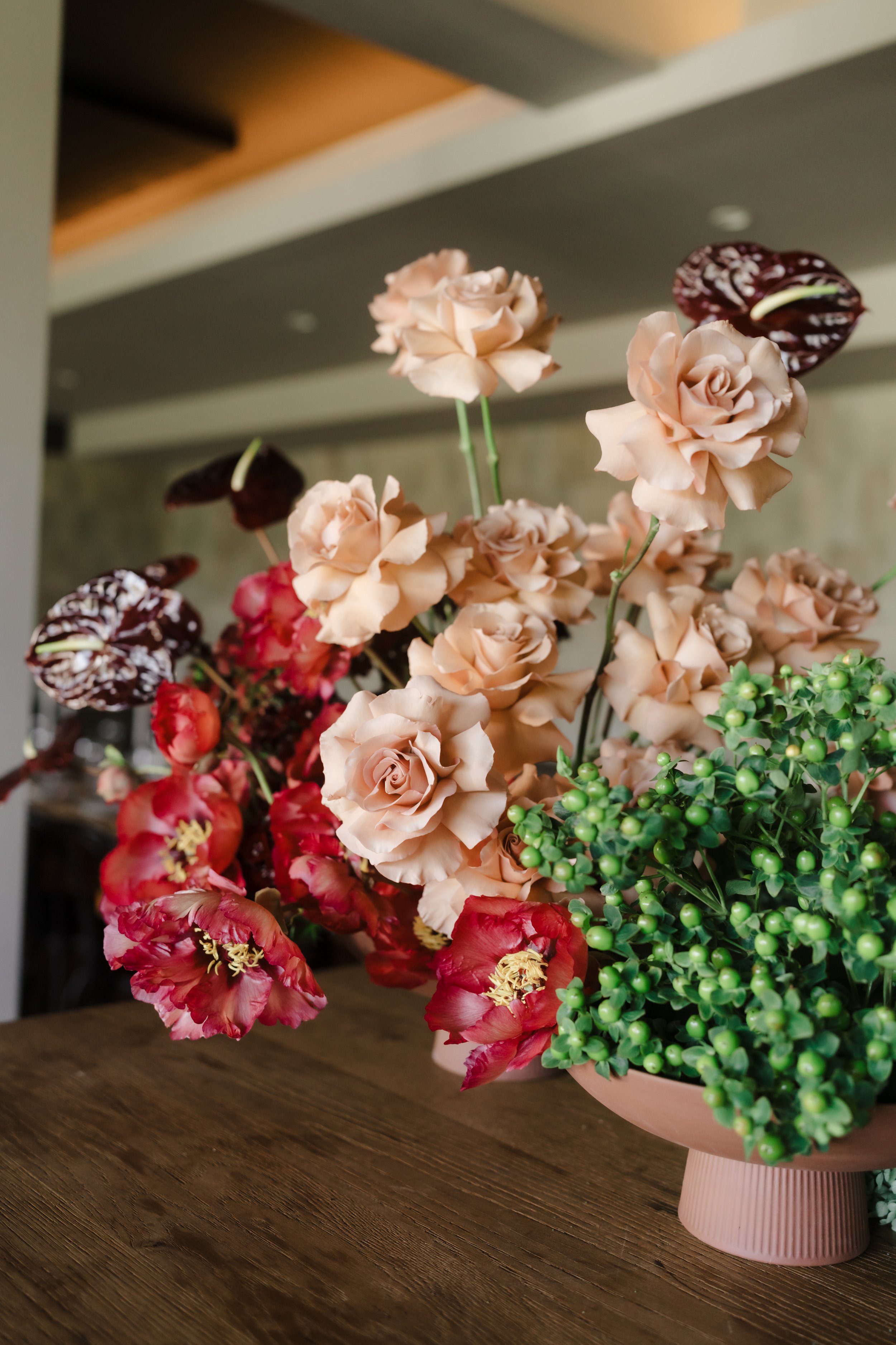 A bouquet of pink roses, red peonies with yellow centers, and dark red anthuriums with white veins, arranged in a pink ceramic vase on a wooden table.