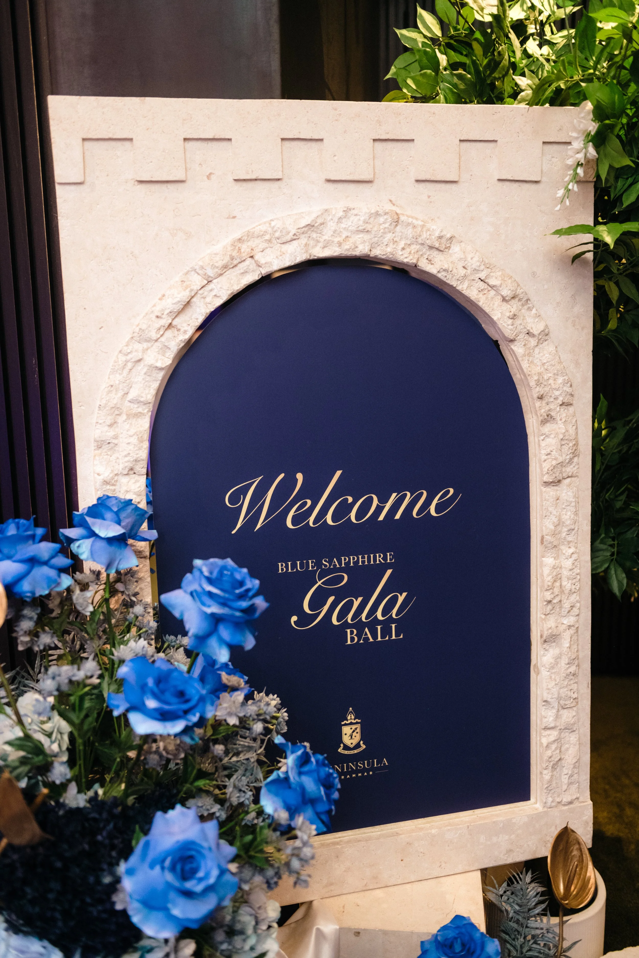 Modern‑lux corporate event styling by Poppy Culture at RACV Cape Schanck Resort, featuring a castle‑style welcome sign for the Blue Sapphire Gala Ball. The deep‑blue panel is framed by a stone‑textured arch and surrounded by blue roses and lush green