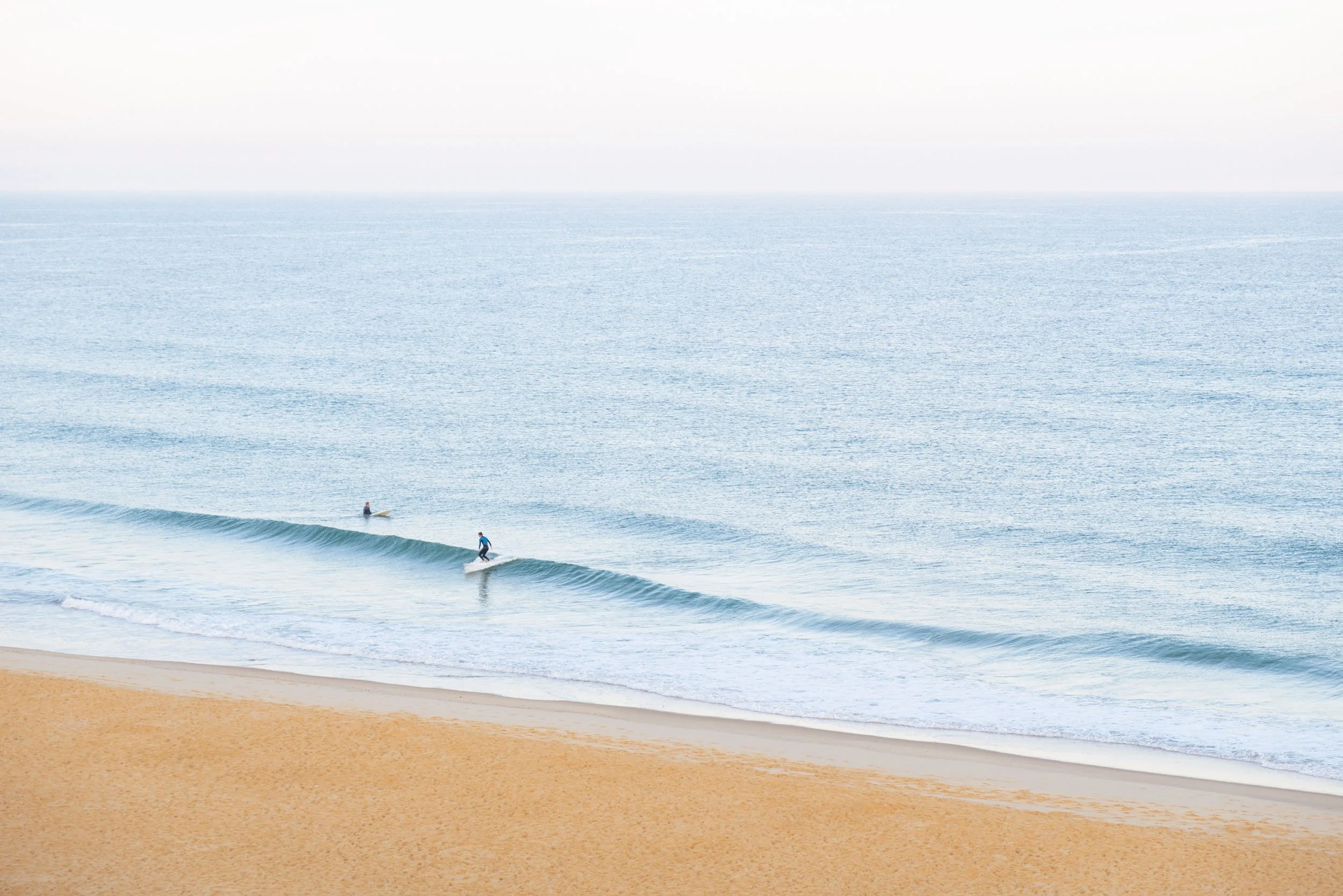 Der Strand mit sanften Wellen, zwei Surfer auf dem Wasser, Himmel ist leicht bewölkt, ruhige Atmosphäre.