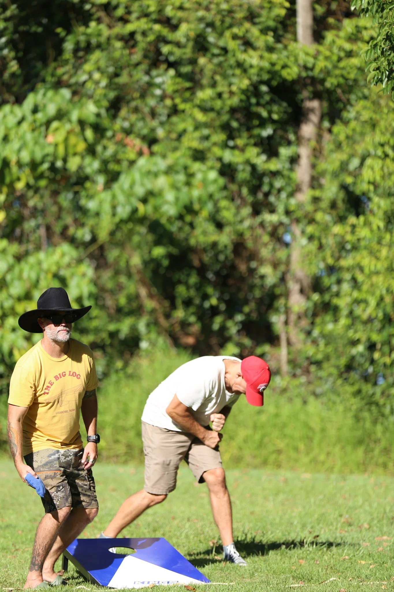 Two men outdoors playing cornhole, one in a yellow T-shirt, camouflage shorts, large black hat, and sunglasses, holding a blue bean bag, the other in a white T-shirt, gray shorts, red cap, crouched down preparing to throw a bean bag, with a cornhole 
