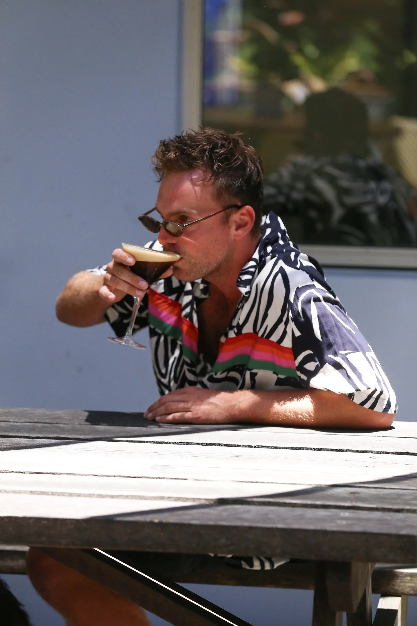 A man with brown hair and sunglasses, wearing a colorful patterned shirt, is sitting at an outdoor table, drinking a dark iced coffee or cocktail.