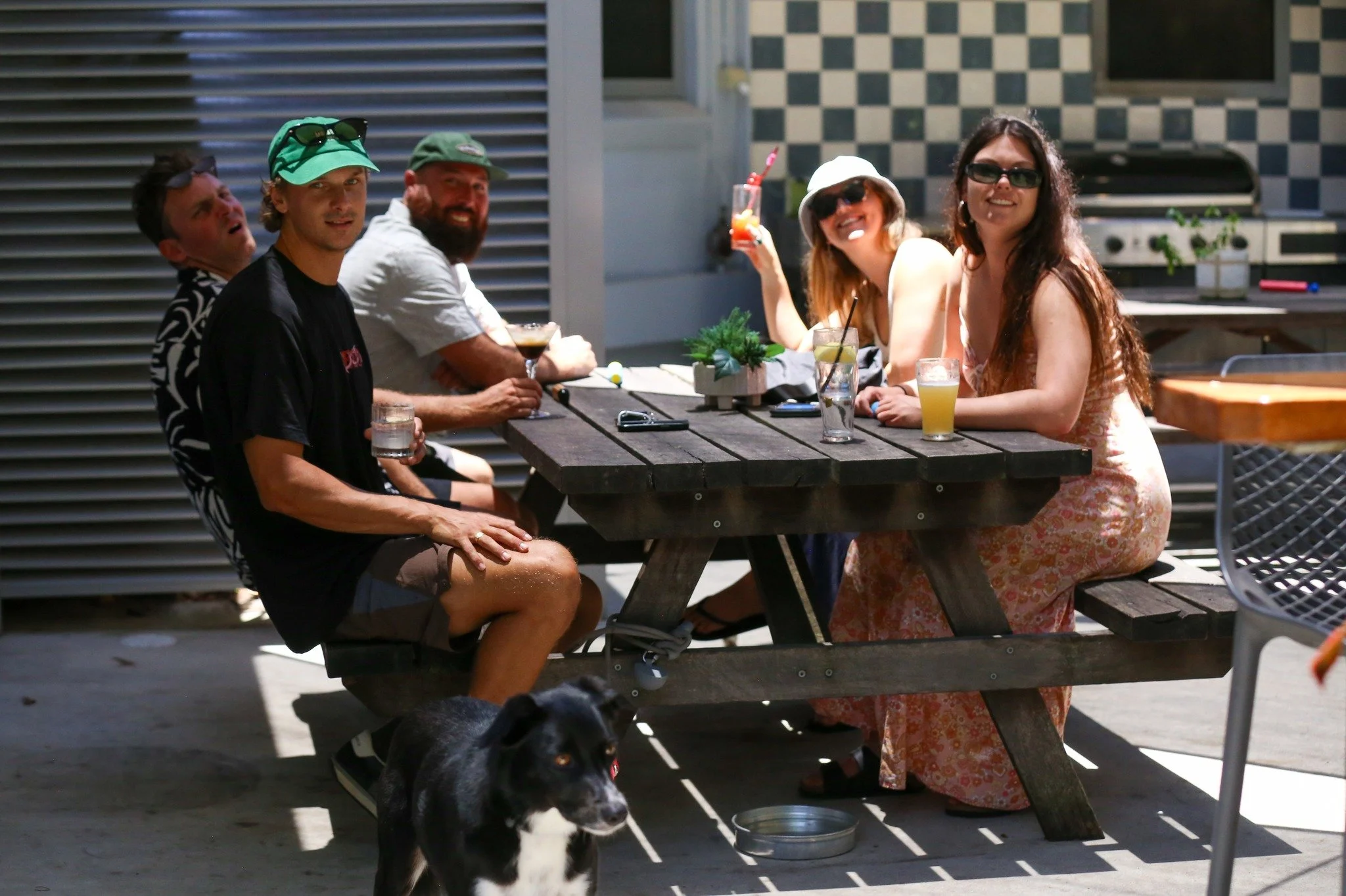 Group of people sitting at a picnic table outdoors, smiling and enjoying drinks; a dog sitting nearby.