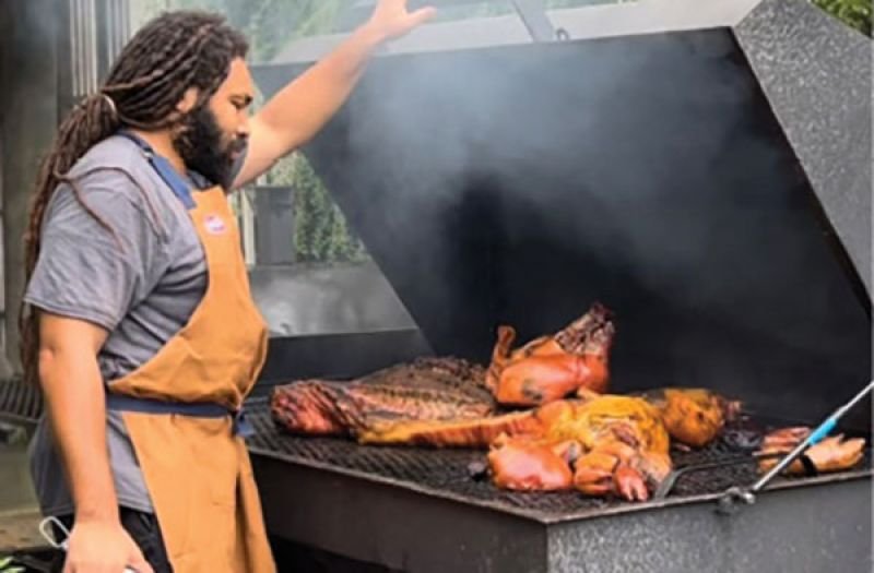Person cooking whole pigs on a large outdoor grill, wearing a brown apron, with smoke rising from the grill.